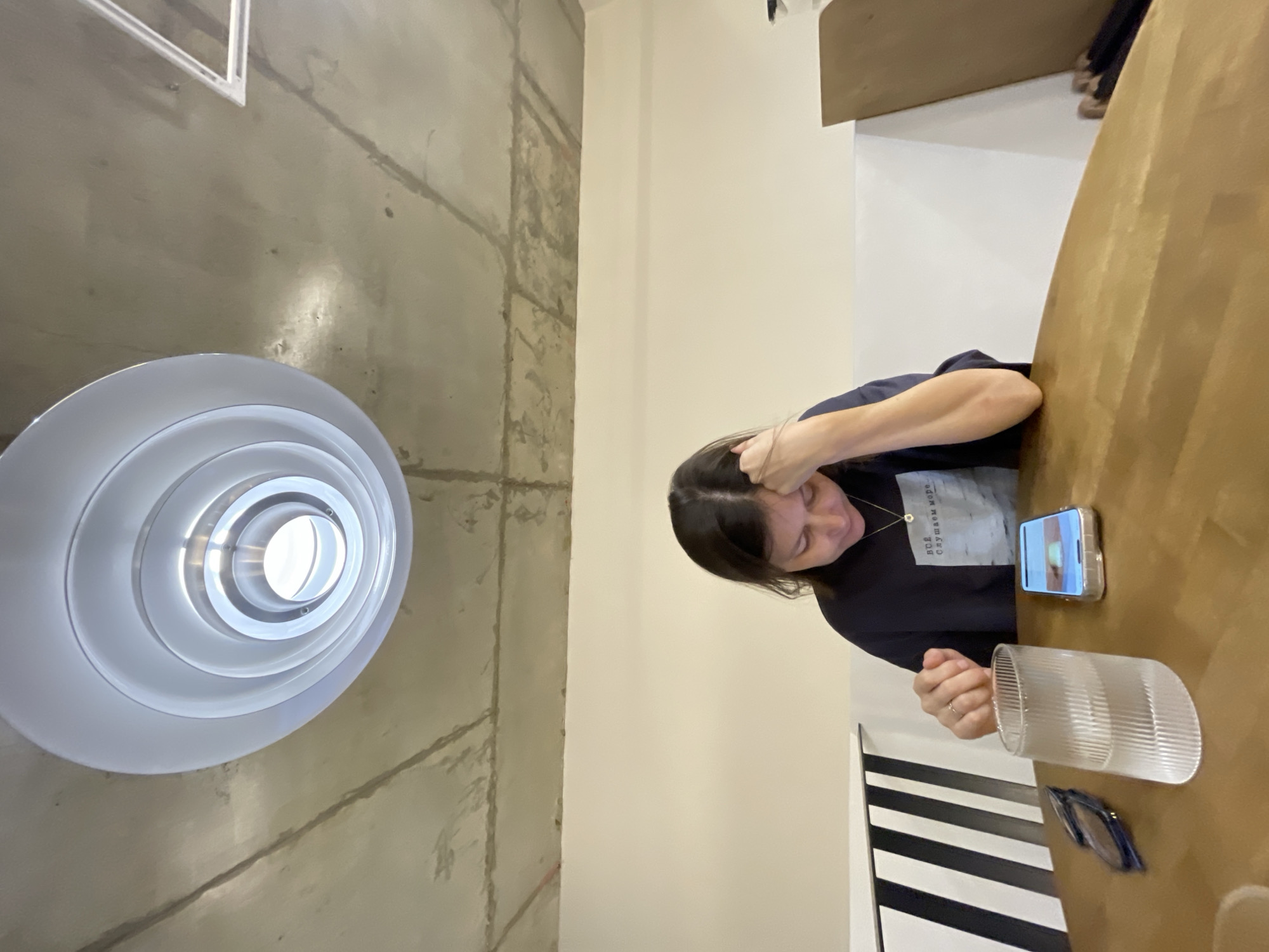 A young woman is seated at a wooden table in a cafe, looking down at her smartphone. She is wearing a dark blue sweatshirt with a graphic print and a necklace. On the table in front of her are the smartphone, a clear ribbed glass with water, and a pair of sunglasses. The cafe has an exposed concrete ceiling with a circular light fixture. The walls are painted white and beige, and a section of the wall features black and white diagonal stripes. The scene is well-lit by overhead lighting. There are no visible indications of the specific location within Izhevsk, Russia, beyond it being a cafe.