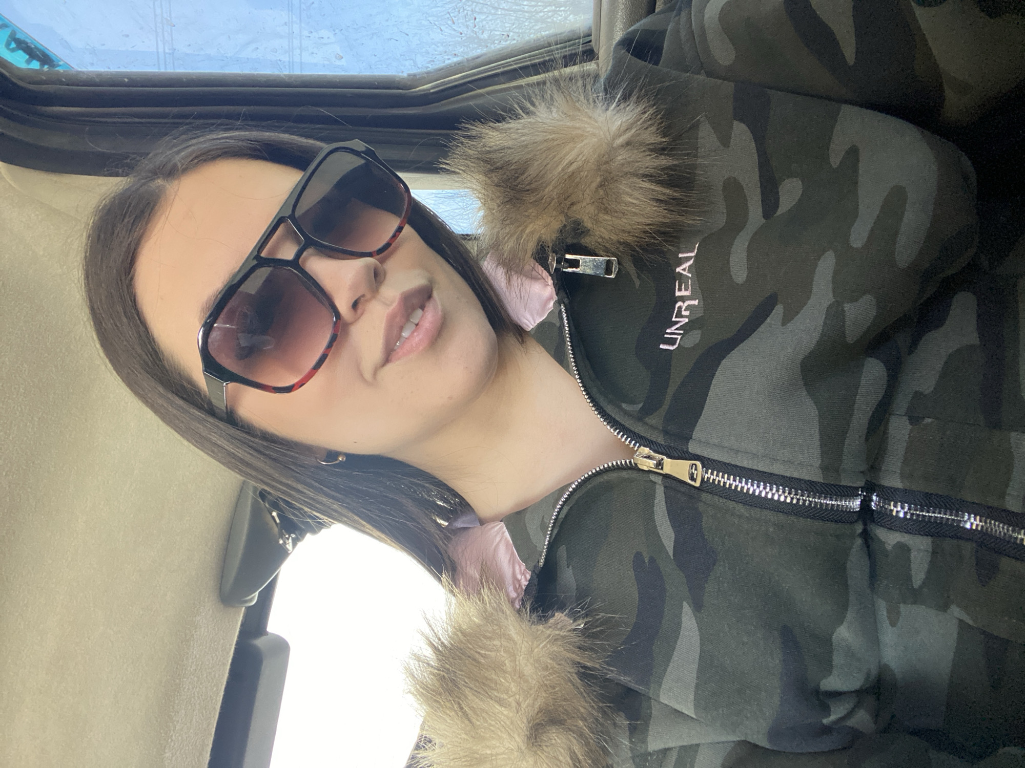 A woman is in a car, likely the driver or front passenger, wearing a camouflage jacket with fur trim and the word "UNREAL" visible on the upper left. She is also wearing sunglasses with a brown/red gradient and a black frame. The interior of the car is beige. The location cannot be determined to be Chaikovsky, Russia based on the visual information provided.