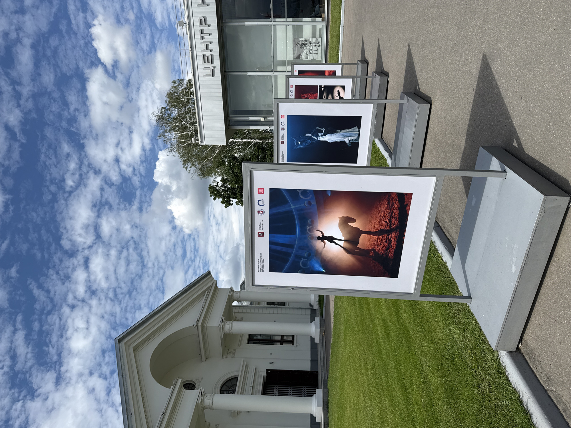 An outdoor exhibition of circus-themed photographs is displayed in front of a building with classical architecture, likely a museum or cultural center, in Moscow, Russia. The building on the left features a white facade with large columns and an arched entrance. To the right, a modern building with large glass windows displays signage that reads "ЦЕНТР Н" in Cyrillic, indicating "Center". Several framed photographs are mounted on stands, arranged in a row on an asphalt pathway. The foreground features a large print of a performer standing on a horse under dramatic lighting. Subsequent photographs show a performer with a dark horse, and other circus-related imagery. The ground is covered with green grass and the sky is blue with scattered white clouds. The overall setting suggests an artistic event or exhibition open to the public.