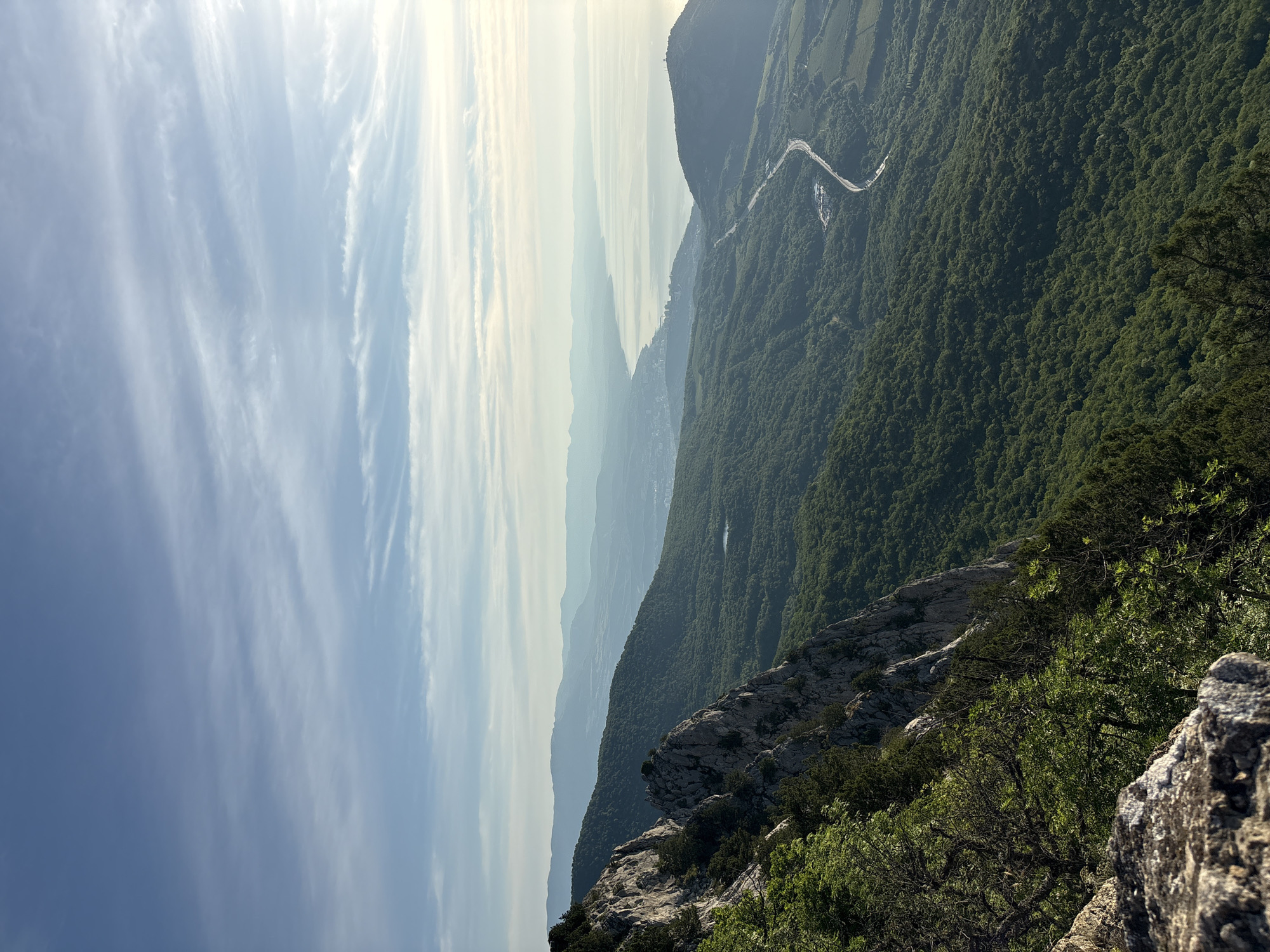 This is an expansive, elevated panoramic view of a mountainous coastal landscape in Malyi Mayak, Ukraine. The upper third of the image is occupied by a light blue sky with scattered wispy cirrus and stratocumulus clouds, gradually brightening towards the right, indicating the sun's position.
In the immediate foreground on the lower left, weathered grey rock formations are visible, partially covered by dark green shrubs and trees. The midground features steep, rugged, grey rock cliffs and rolling hills, densely covered in a mixed forest of dark green trees and shrubs. A winding asphalt road is discernible in the lower-mid right, cutting through the forest, with a small white building located at one of its bends. A small, dark, circular body of water, likely a lake or pond, is situated within the forested hills in the mid-left portion of the image.
Further into the background, successive layers of mountains recede, their contours softened by atmospheric haze. The coastline of the Black Sea stretches horizontally across the right midground, reflecting the sky's brightness. Along this coastline, an urbanized area, representative of Malyi Mayak and surrounding settlements, is visible with numerous buildings. A communications tower or similar structure stands prominently on a hill near the coast on the far right. The scene is illuminated by natural sunlight from the right, casting soft shadows and highlighting the varied terrain, consistent with morning or late afternoon light conditions. No human figures or animals are visible.