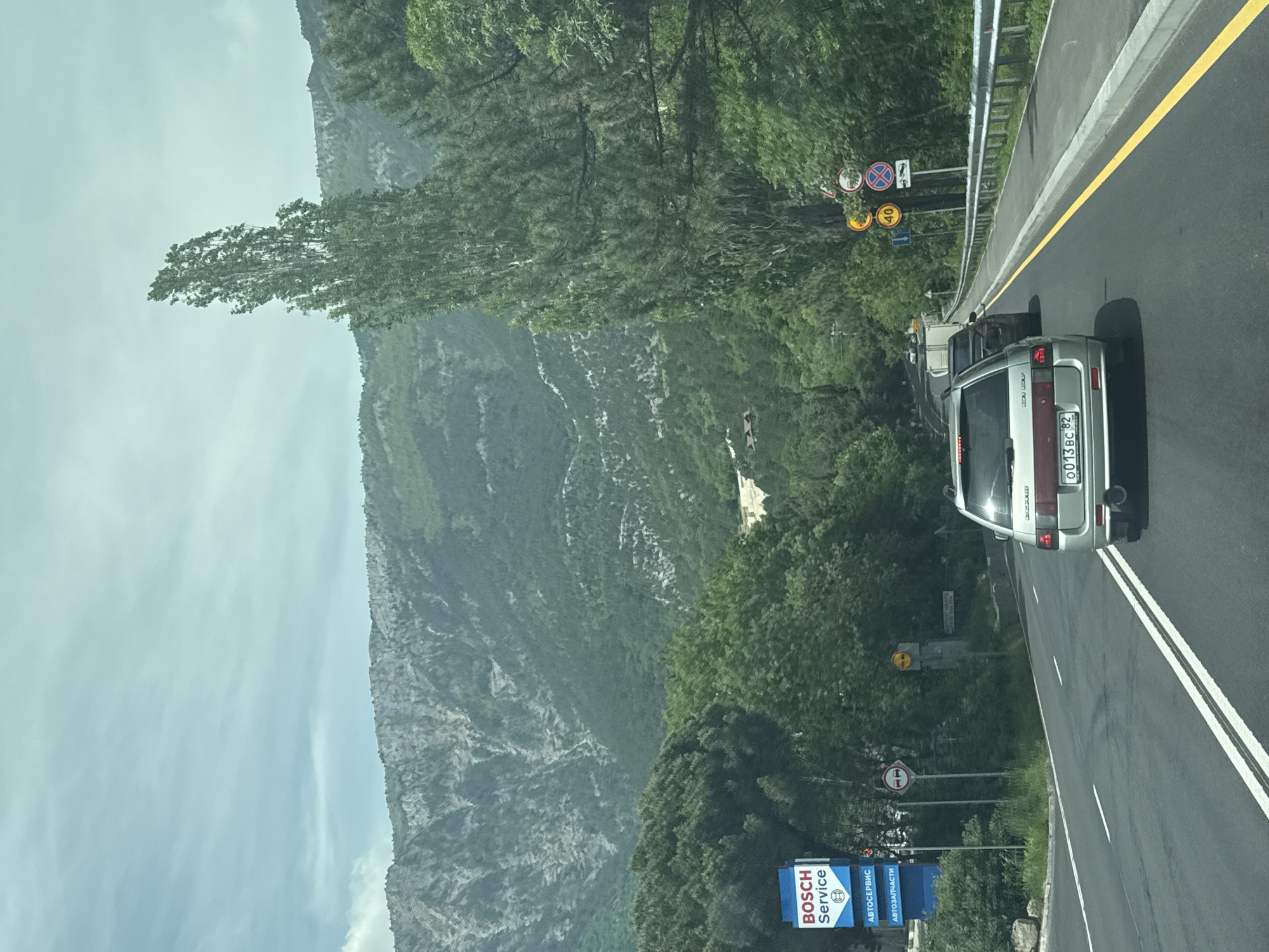 The image depicts a road in the vicinity of Ливадийский поселковый совет, Ukraine, featuring a gray car with the license plate "0013 BC 82" driving on the right lane. The road has a yellow line along the right edge and double white lines separating lanes. A metal guardrail runs along the side of the road. Traffic signs are visible on the left side of the road, along with a "Bosch Service" sign. The background consists of a mountainside covered in trees and vegetation. The sky is partly cloudy.