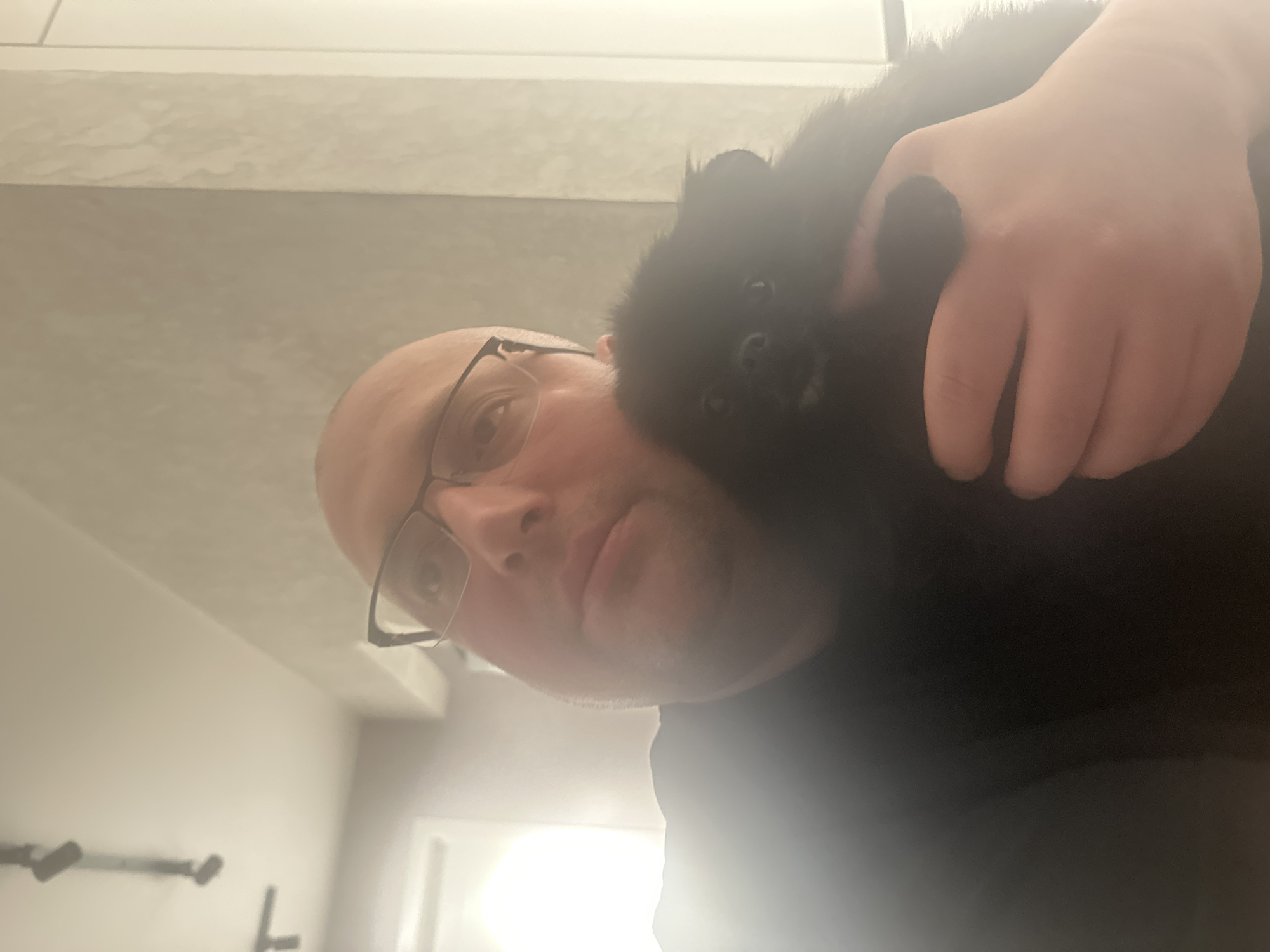 A man wearing glasses holds a small, black dog. The background appears to be a domestic interior. There is no visible evidence to suggest a location within the city of Izhevsk, Russia.