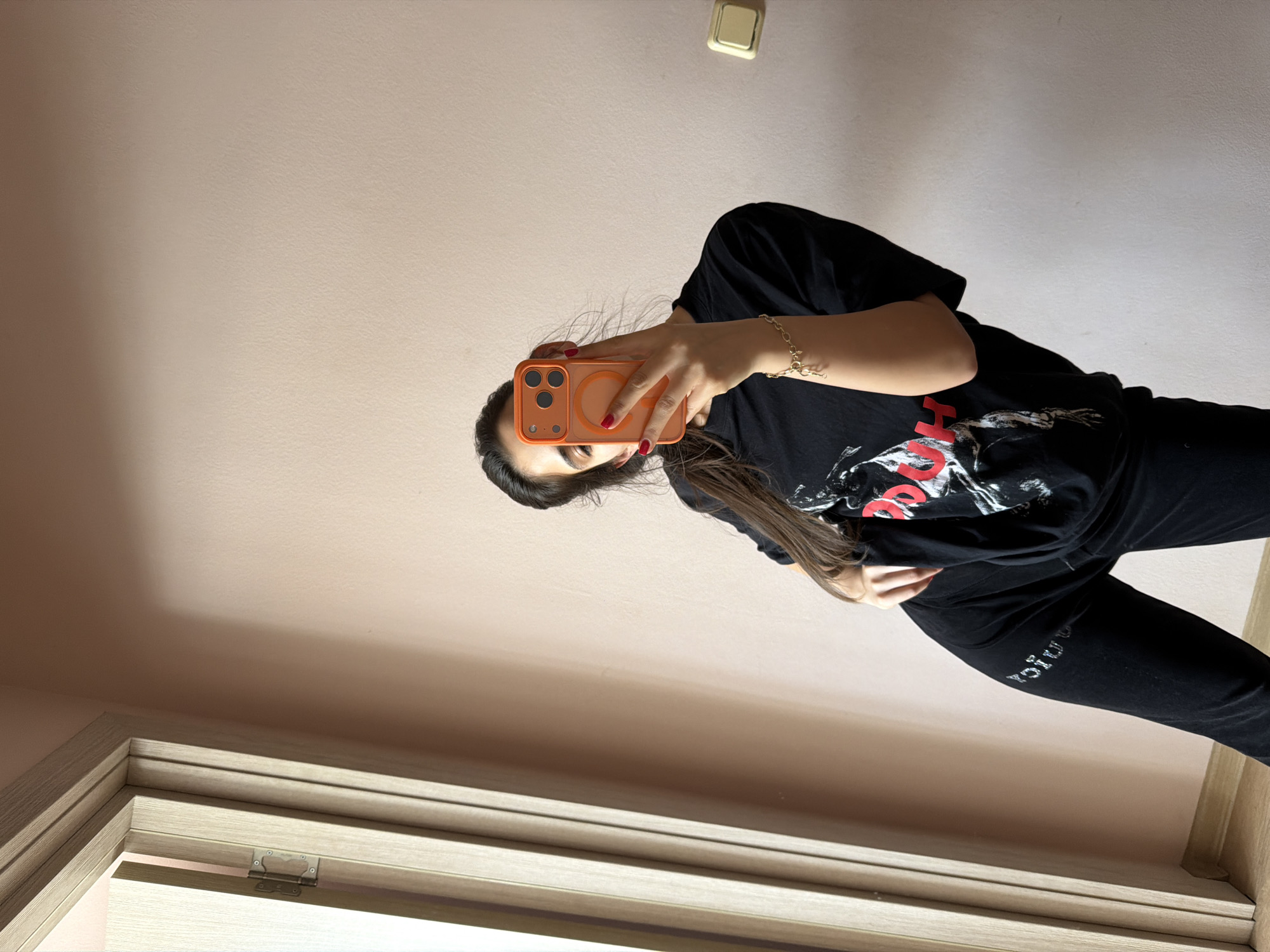 A woman is taking a selfie. She is wearing a black T-shirt and black pants. She is holding an orange smartphone. Her face is partially obscured by the phone. She is wearing a gold bracelet. The photo is taken indoors, possibly in a hallway or room, as evidenced by the door frame. The wall is painted a light peach color. A light switch is visible on the wall. The location is likely in Rudartsi, Bulgaria, based on the prompt's request.