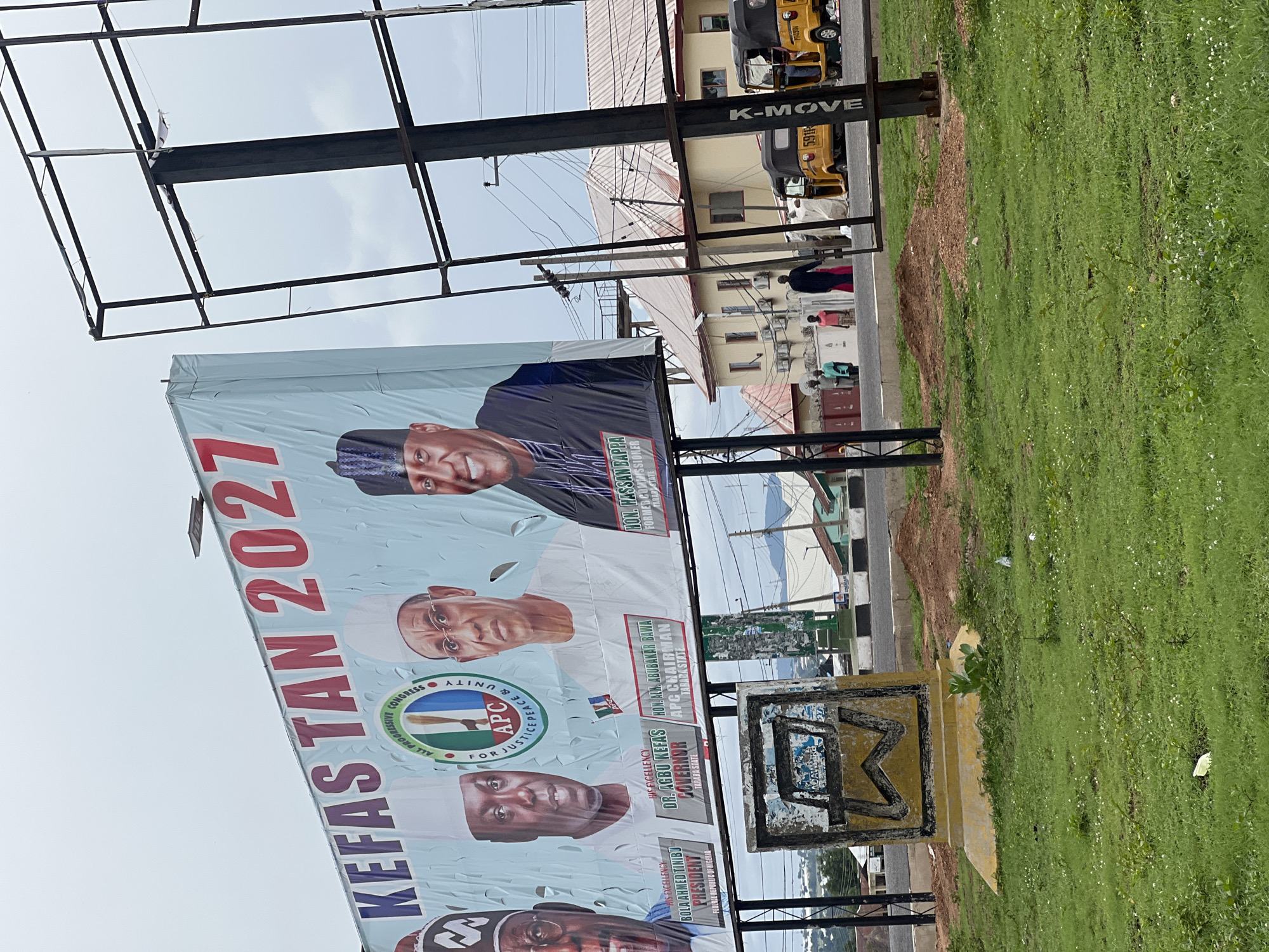 In Jalingo, Nigeria, a large billboard announces "KEFAS TAN 2027" and features portraits of four men. From left to right, the men are identified as "HIS EXCELLENCY BOLA AHMED TINUBU PRESIDENT FEDERAL REPUBLIC OF NIGERIA," "HIS EXCELLENCY DR. AGBU KEFAS GOVERNOR TARABA STATE," "HON. ABUBAKAR BAWA APC CHAIRMAN TARABA STATE," and "HON. HASSAN SANTA FORMER COMMISSIONER TARABA STATE." The billboard also displays the logo of the All Progressives Congress (APC) party. The billboard is weathered and shows signs of wear. In the foreground, a grassy area with some scattered debris is visible. To the right of the billboard, a metal frame for another billboard stands empty. In the background, buildings, power lines, and people can be seen. Several yellow auto-rickshaws are also present on the road.