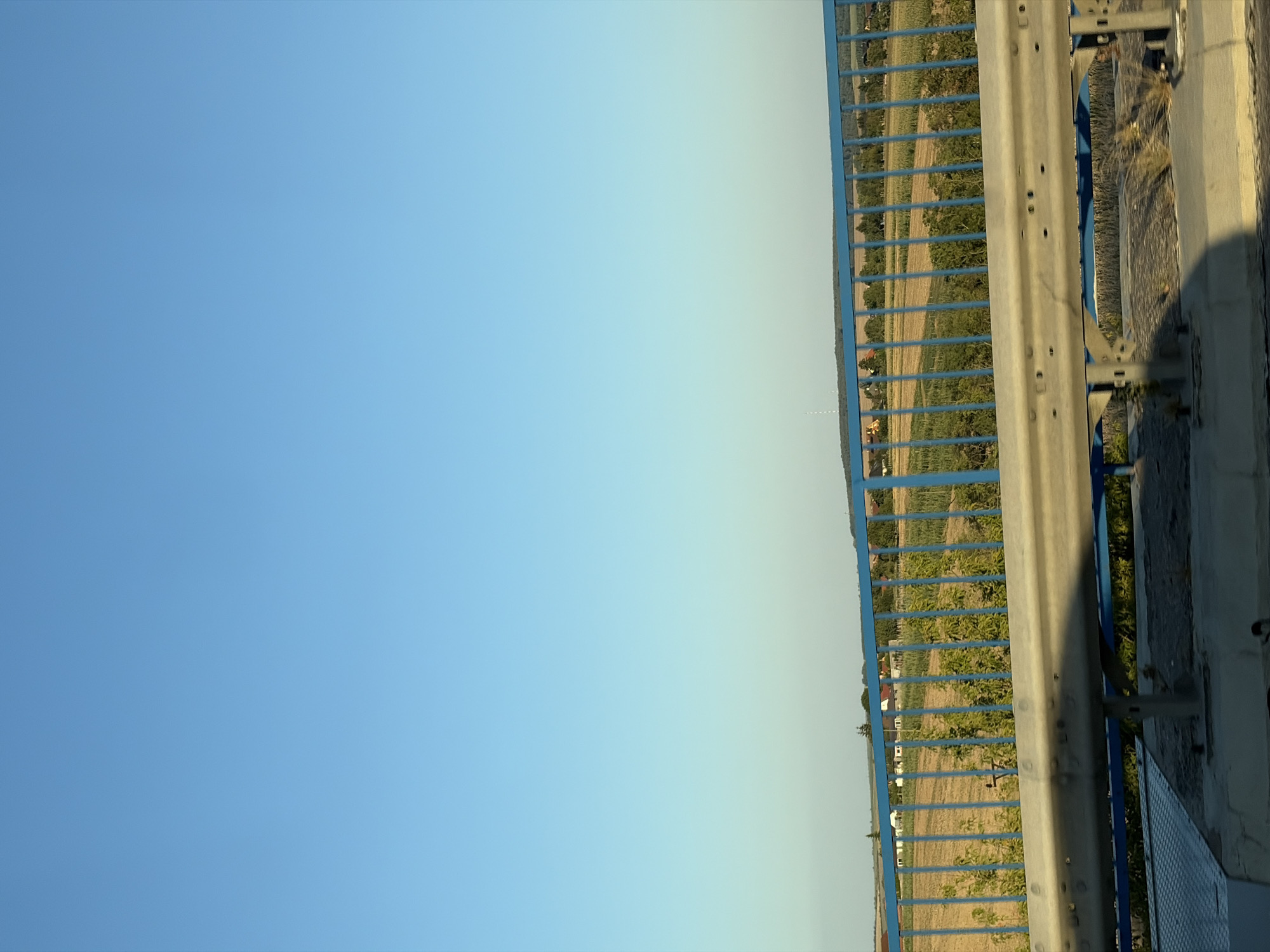 The image captures a view from a bridge overlooking a rural landscape in or near Veľké Zálužie, Slovakia. The sky is a clear, gradient blue, transitioning from a lighter hue at the horizon to a deeper shade overhead. A prominent blue metal railing runs across the foreground, separating the viewer from the scene below. Below the railing, a metal guardrail is visible, reflecting sunlight and casting a shadow. Beyond the guardrail, a field of dry grass or harvested crops stretches out, interspersed with rows of green foliage, possibly young trees or shrubs. In the distance, a cluster of houses with red roofs suggests a small village or settlement. The overall environment is serene and agricultural, under a vast expanse of sky.