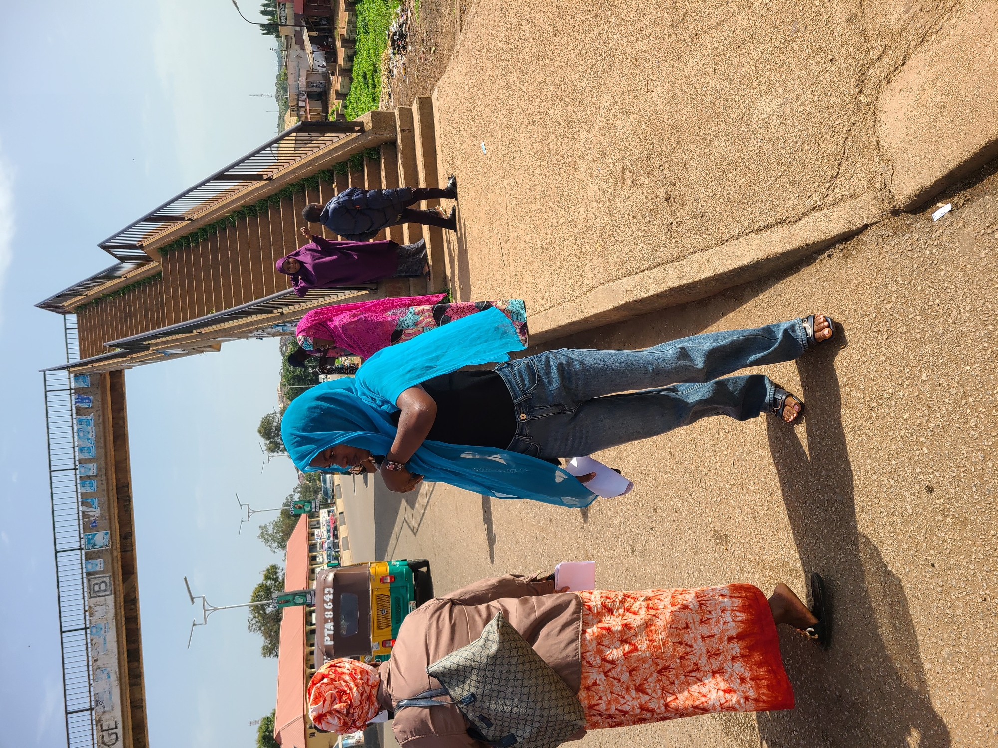 The image depicts a street scene in Gura Top, Nigeria, with a focus on a pedestrian overpass and several individuals. In the foreground, a woman wearing a bright blue hijab and blue jeans stands with her back partially to the camera, holding a white sheet of paper. To her left, another woman is seen from the back, wearing an orange and white patterned skirt, a brown jacket, and a patterned headscarf. She carries a large, patterned tote bag over her shoulder and also holds a white paper. A three-wheeled auto-rickshaw with the license plate "PTA-8643" is visible behind them.
The pedestrian overpass dominates the background, with a wide staircase leading upwards. Several people are on or near the stairs: a woman in a bright pink headscarf and patterned dress, another woman in a dark purple abaya, and a young person wearing a dark jacket and trousers. The overpass itself appears weathered, with posters or signs affixed to its underside. The surrounding environment includes buildings, streetlights, and vegetation, suggesting an urban setting. The ground is paved with asphalt and concrete. The lighting indicates a daytime scene, likely under a bright sky with some clouds.