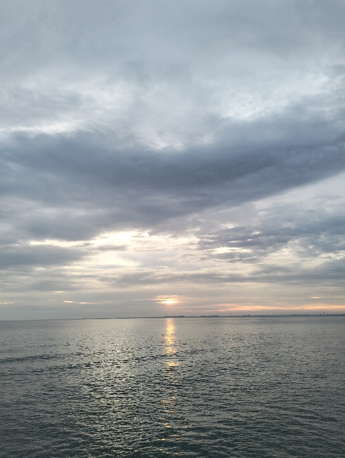 The image depicts a serene seascape under an overcast sky. The water is calm with subtle ripples and a shimmering path of light reflecting the setting or rising sun. The sky is dominated by layers of grey and white clouds, obscuring the sun except for a faint glow on the horizon. A distant coastline or landmass is barely visible across the water, adding depth to the scene. The overall mood is peaceful and tranquil.