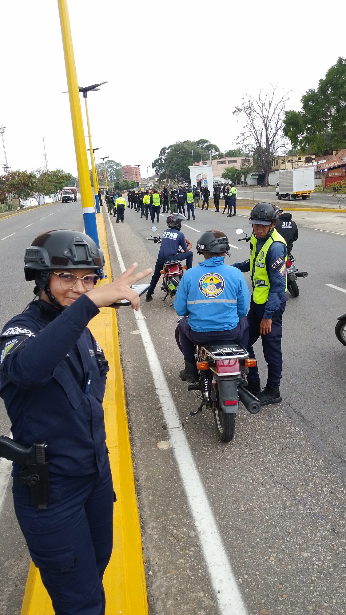 A medium shot depicts a group of law enforcement officers on a road in Tinaquillo, Venezuela. A uniformed officer, equipped with a helmet and firearm, stands near a yellow and blue curb on the road's edge. She is wearing glasses and giving the peace sign, looking at the viewer. Several officers on motorcycles are nearby; two are pulled over on the side of the road and one is talking to a person sitting on their motorcycle. In the background, a line of additional officers is present. The area appears to be on the outskirts of the city, with buildings and trees visible in the distance. A large white building is slightly out of focus in the background to the right.