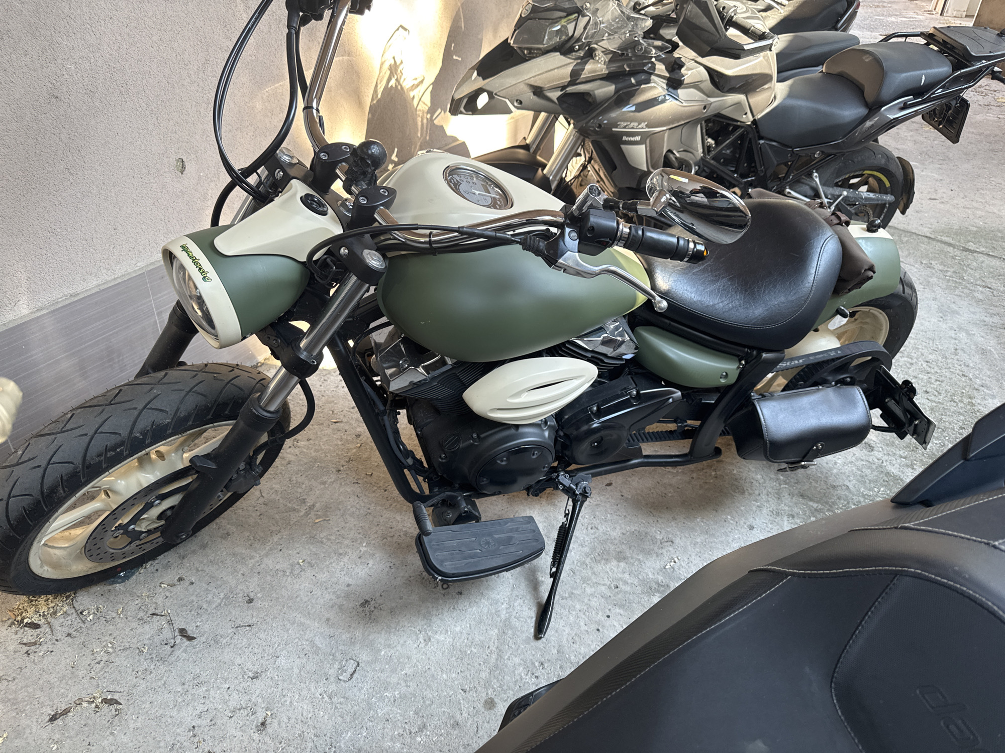 The image shows a parked green and cream-colored motorcycle with black accents. It has a wide front tire and a black seat. A black leather saddlebag is mounted on the rear of the motorcycle. Behind the first motorcycle, another motorcycle is partially visible. The motorcycles are parked on a concrete surface, possibly a garage or parking area, adjacent to a light-colored wall. The location is indicated to be in the city of Plovdiv, Bulgaria.