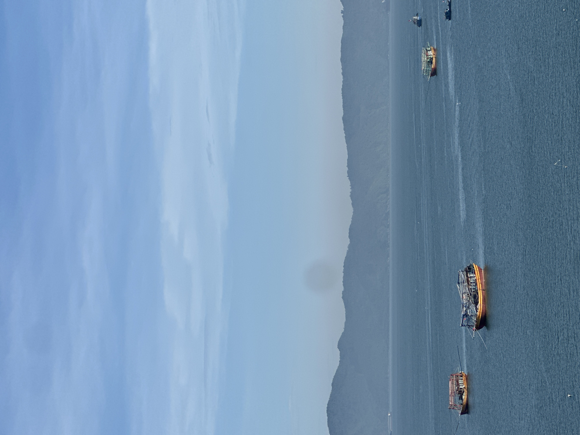 The image depicts a serene seascape with several boats on the water, set against a backdrop of lush, hazy mountains. The sky is a soft blue with wispy clouds, suggesting it is daytime. In the foreground, the deep blue water is calm, with gentle ripples. Two fishing boats are prominently featured in the mid-left of the frame, appearing to be anchored or drifting. They have distinct hulls, with one being predominantly yellow and red, and the other a mix of red and yellow, both covered with a protective structure. Further out in the water, more boats are visible, including a larger, covered boat in the center and several smaller vessels in the distance. The mountains in the background are a muted green and blue, shrouded in a light mist, which adds to the tranquil atmosphere. This scene is consistent with views from the Son Tra Peninsula in Da Nang, Vietnam, looking out over the East Sea. The overall impression is one of peace and the natural beauty of the coastal landscape.