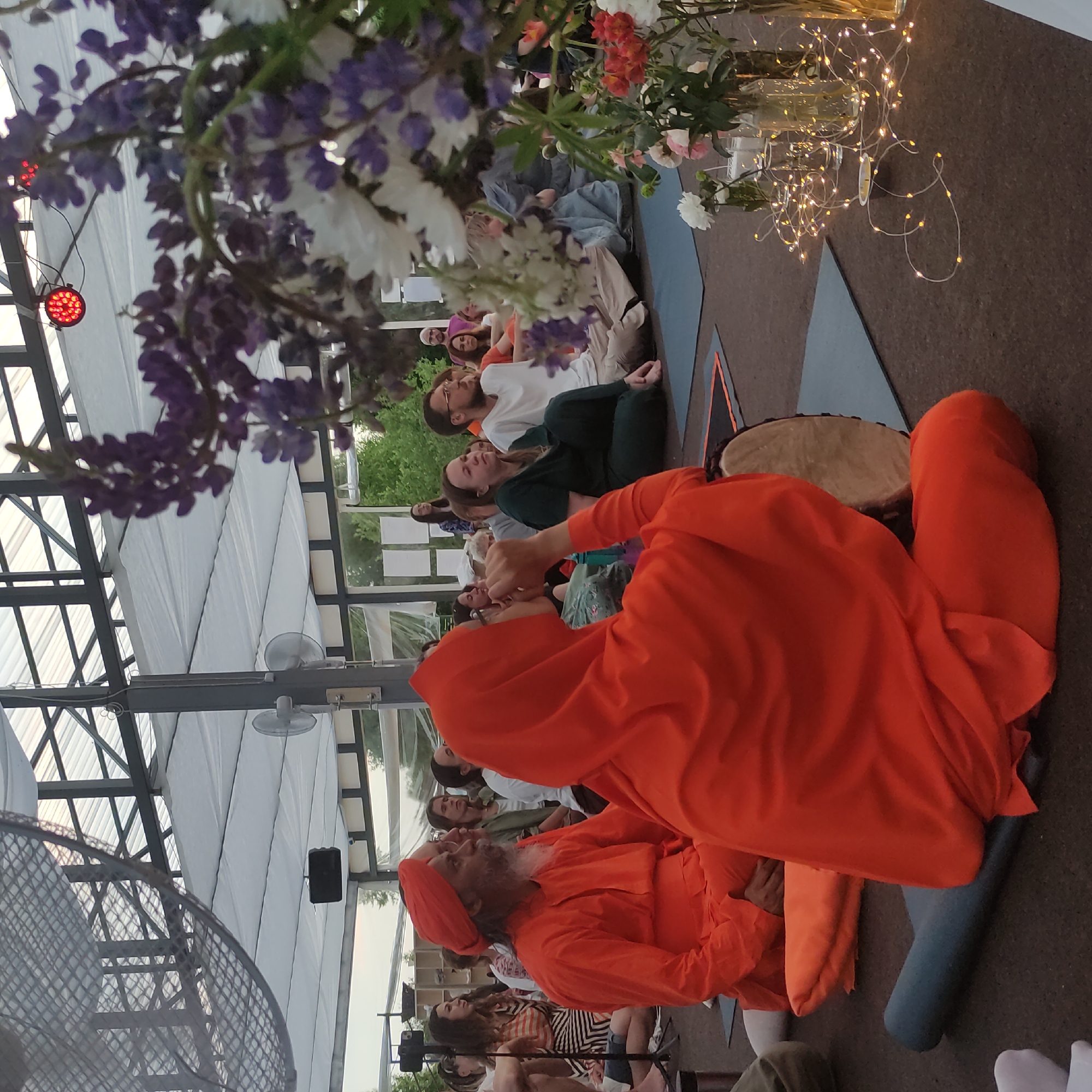 The image captures an outdoor gathering under a translucent canopy. In the foreground, two individuals are prominently featured. One, on the left, is a man wearing an orange turban and robe, with a beard, sitting cross-legged and holding a drum. Beside him, a person in an orange robe is also seated cross-legged, facing away from the camera, with their hands clasped in front of their face. The background reveals a diverse group of people sitting on the floor, some facing the man in the turban, suggesting an event or ceremony. The environment includes decorative elements like flowers in vases and string lights, contributing to a festive or spiritual atmosphere. The location is identified as Zhirnosovo, Russia.