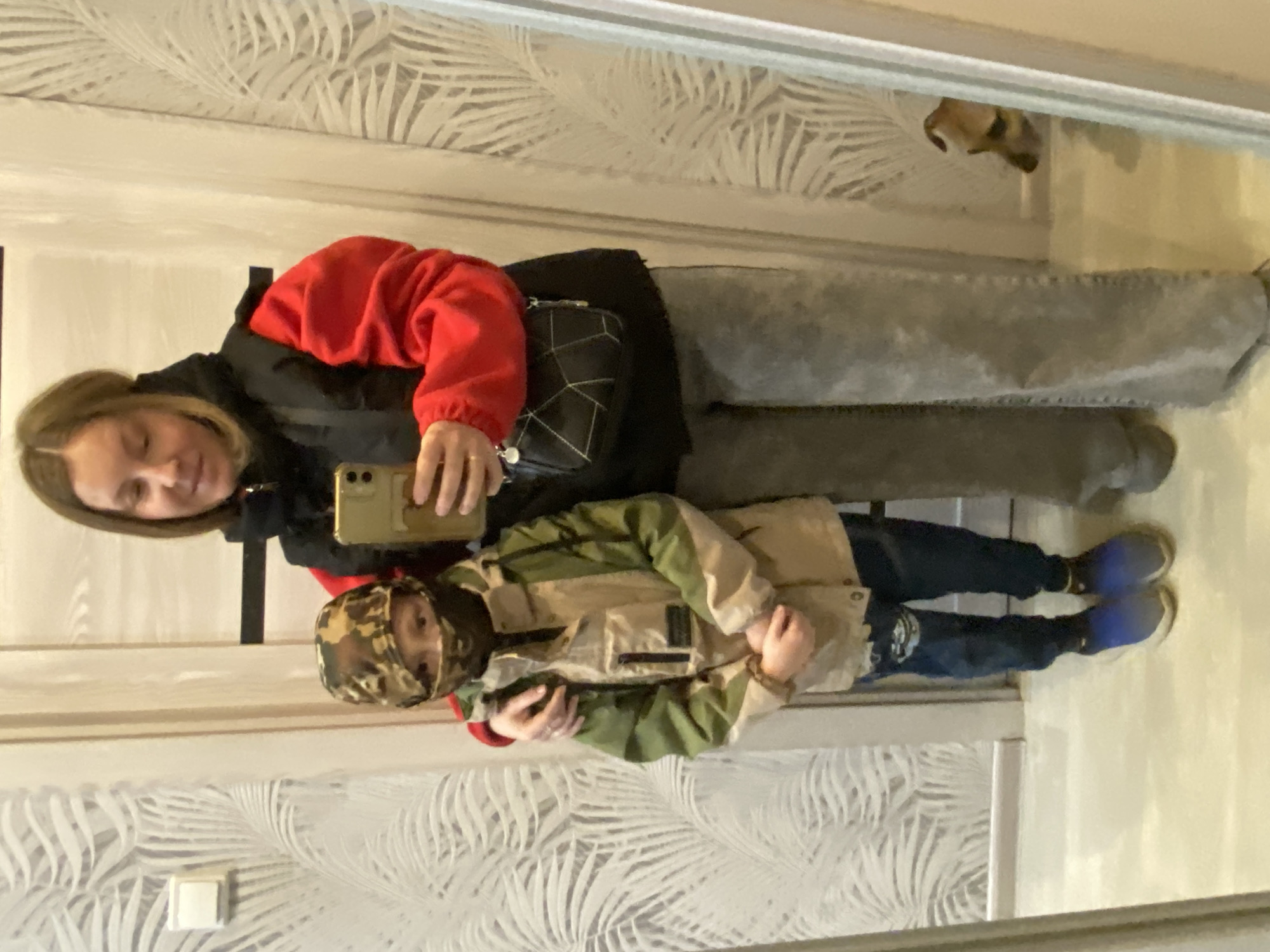 A woman and a child are standing in front of a mirror. The woman is wearing a red and black jacket, gray pants, and is holding a cell phone. The child is wearing a camouflage hat, a khaki and green jacket, jeans, and blue shoes. They are posing for a photo in what appears to be an apartment with patterned wallpaper. The image is taken in Noviy, Russia. A dog is visible in the mirror.