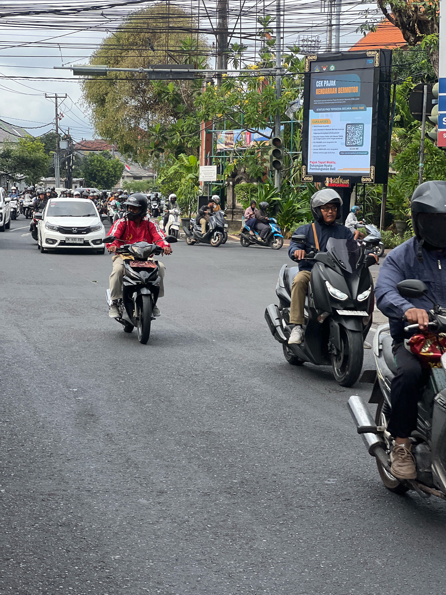 This image depicts a busy street scene in Denpasar, Indonesia. The focus is on a traffic intersection with numerous motorcycles and a few cars navigating the road. Overhead, a dense network of electrical wires crisscrosses the sky.
In the foreground, a man wearing a red jacket and helmet rides a black motorcycle, heading towards the viewer. To his right, another man, also wearing a helmet and glasses, is on a black maxi-scooter, also moving forward. Further to the right, the back of a third motorcyclist is visible, with their motorcycle's exhaust pipe prominent.
In the background, more motorcycles and a white car are visible. A large billboard advertising "CEK PAJAK KENDARAAN BERMOTOR" (Check Motorcycle Vehicle Tax) stands to the right, featuring a QR code and information in Indonesian. Traffic lights are also present, indicating this is an active intersection. Lush green trees and vegetation line the sides of the road, with glimpses of buildings and rooftops behind them. The overall atmosphere suggests a typical day of urban traffic in the city.
