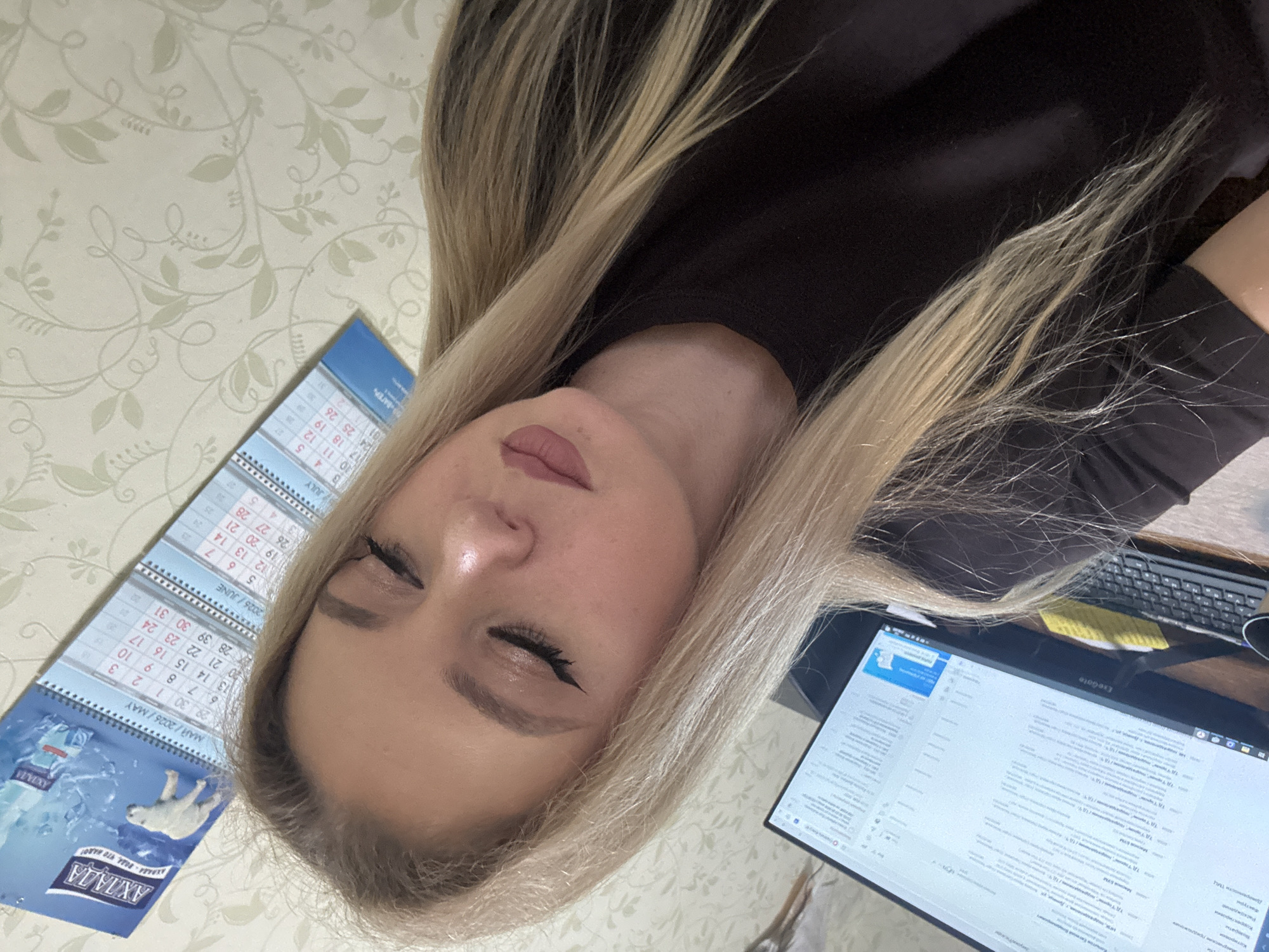 A woman with blonde hair is captured in a close-up selfie, her eyes closed, with makeup applied to her face. She is wearing a black t-shirt. In the background, a computer monitor displays what appears to be an email interface or a similar digital document. To the right, a calendar is visible, showing the months of May, June, and July, with "MAЙ/2026/MAY" clearly legible. The wall behind her is adorned with a light-colored floral wallpaper. The scene suggests an indoor setting, possibly an office or a home workspace.