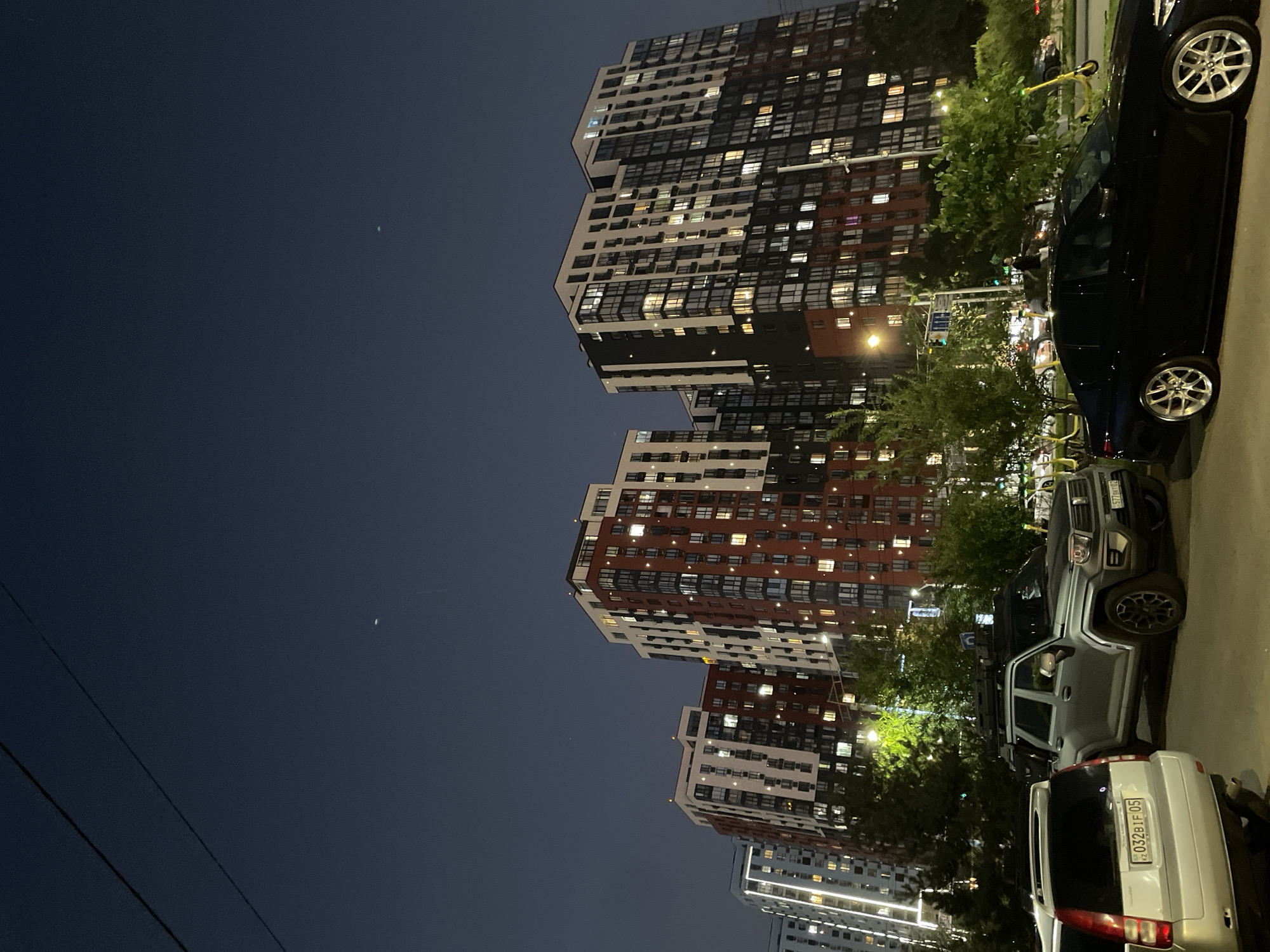 This is a nighttime photograph taken in the city of Almaty, Kazakhstan. The scene depicts a row of modern, multi-story apartment buildings with many illuminated windows, suggesting it's an urban residential area. In the foreground, several vehicles are parked: a silver SUV on the left, a gray off-road vehicle in the middle, and a black sedan on the right. Trees and streetlights are visible, adding to the urban nighttime ambiance. The sky is dark with a few distant lights, possibly stars or distant aircraft. The image captures a typical street view of an inhabited residential complex at night in Almaty.