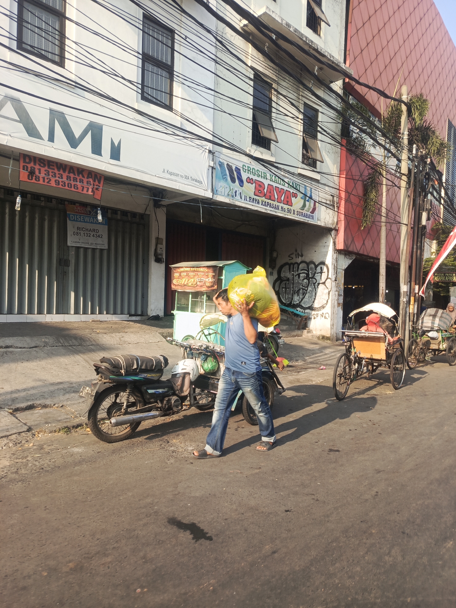 A man in blue jeans and a blue t-shirt carries a large, bright yellow bag of trash over his right shoulder as he walks on a sun-drenched street in Surabaya, Indonesia. The man is walking in front of a parked motorcycle, and behind him are two becaks, or cycle rickshaws, with passengers. The buildings in the background have signs in Indonesian, including "DISWAKAN" (For Rent) and "GROSIR KAOS KAKI 'BAYA'" (Wholesale Socks 'BAYA'). The street appears to be a commercial area, with storefronts and utility poles lining the road. The sky is clear, indicating a sunny day.