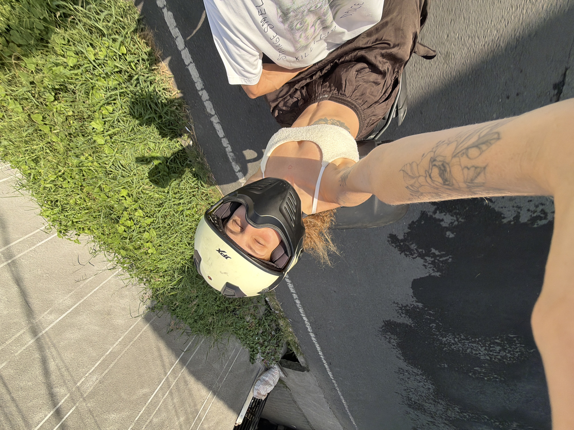 In North Kuta, Indonesia, a POV shot captures two individuals on a scooter, bathed in the golden light of a setting sun. The foreground is dominated by the arm of the person taking the photo, adorned with a floral tattoo. Behind them, a woman with blonde curly hair wears a light-colored helmet and a white crop top, her eyes closed as if enjoying the ride. She is seated behind the driver, whose white t-shirt is visible. They are on a paved road, bordered by lush green grass and a concrete gutter. The scene is characterized by the interplay of light and shadow, with the sun casting long shadows across the road.