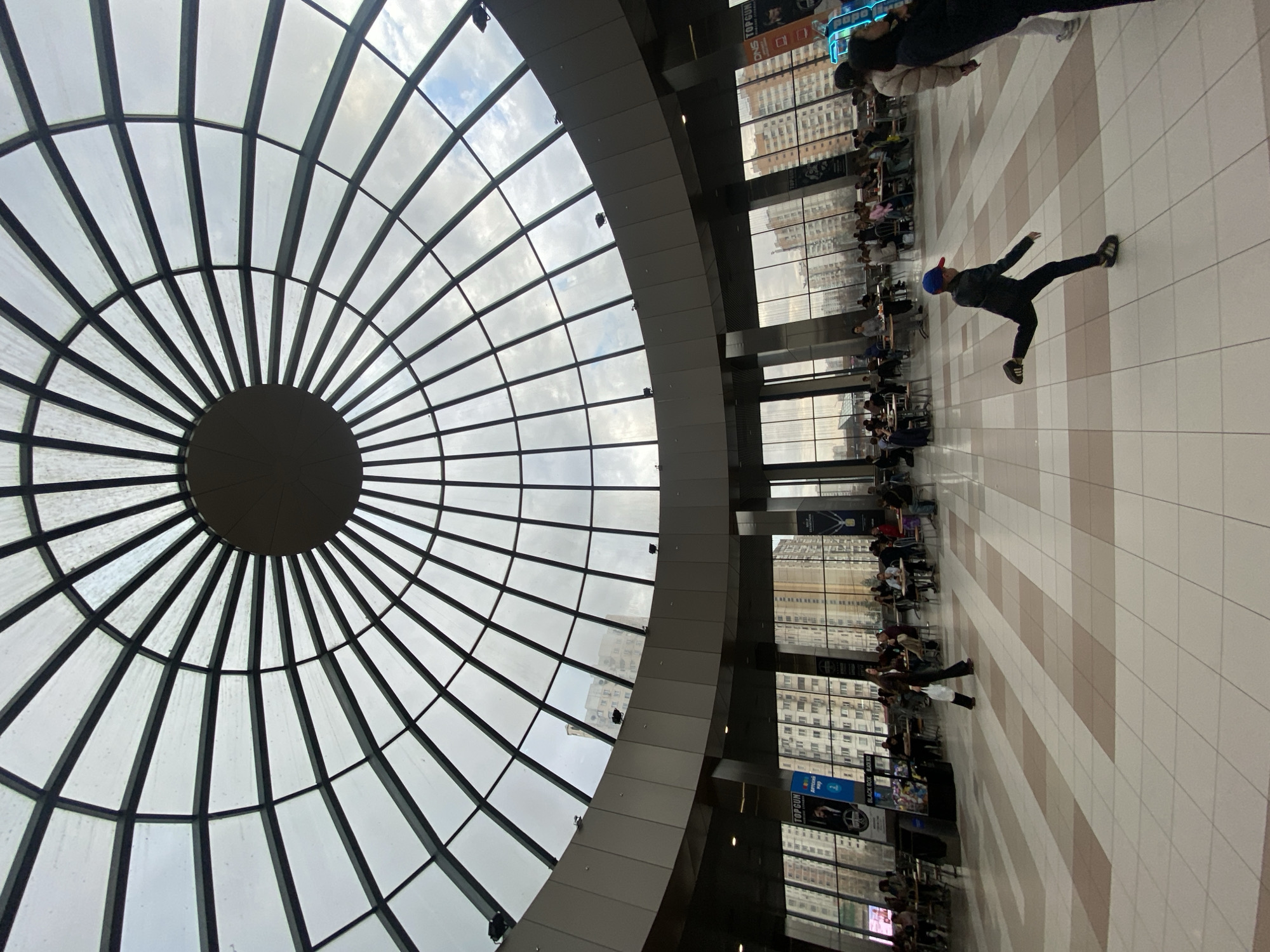 The photograph shows the interior of a building in Moscow, Russia. A large, circular, domed glass ceiling dominates the upper left portion of the image, allowing natural light to filter through. Below the dome, the interior space features a tiled floor with alternating light and dark beige colored tiles. The right portion of the image displays people seated at tables near large windows overlooking a city street with tall buildings. A person in a dark jacket, blue hat, and sneakers is captured mid-motion on the tiled floor. Posters and signage are visible near the windows on the right.