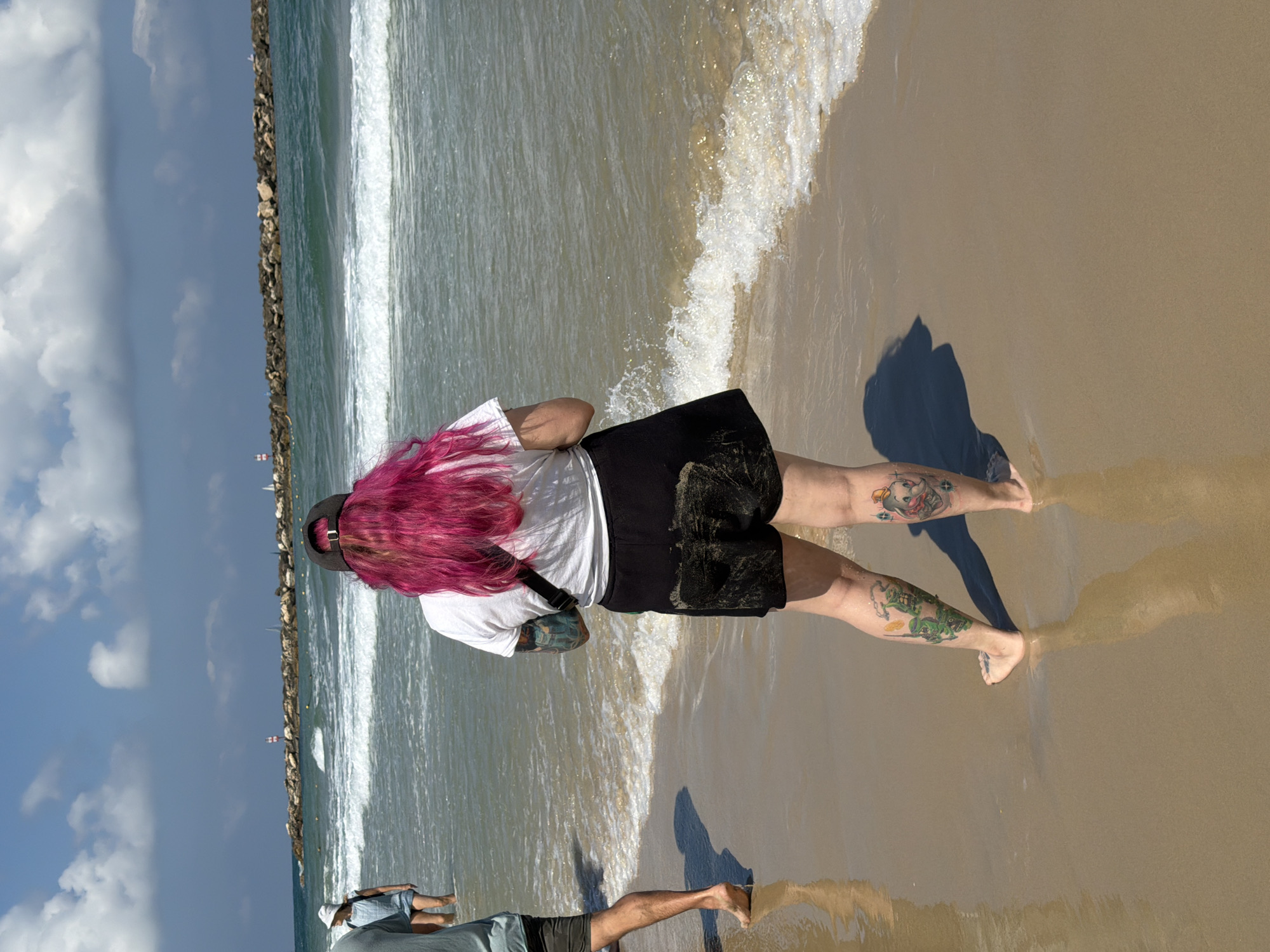 A medium, eye-level shot depicts a person with bright pink hair standing on a beach, with a wave lapping around their feet. The person is wearing a white t-shirt, black shorts, and a black baseball cap. They have tattoos on their legs and arm. A stone breakwater is visible in the background, separating the beach from the Mediterranean Sea. A second person is visible in the lower left corner of the image, also standing on the beach. The sky is blue with scattered clouds. The sand is wet and light brown in color.
The location is a beach in Tel Aviv, Israel.