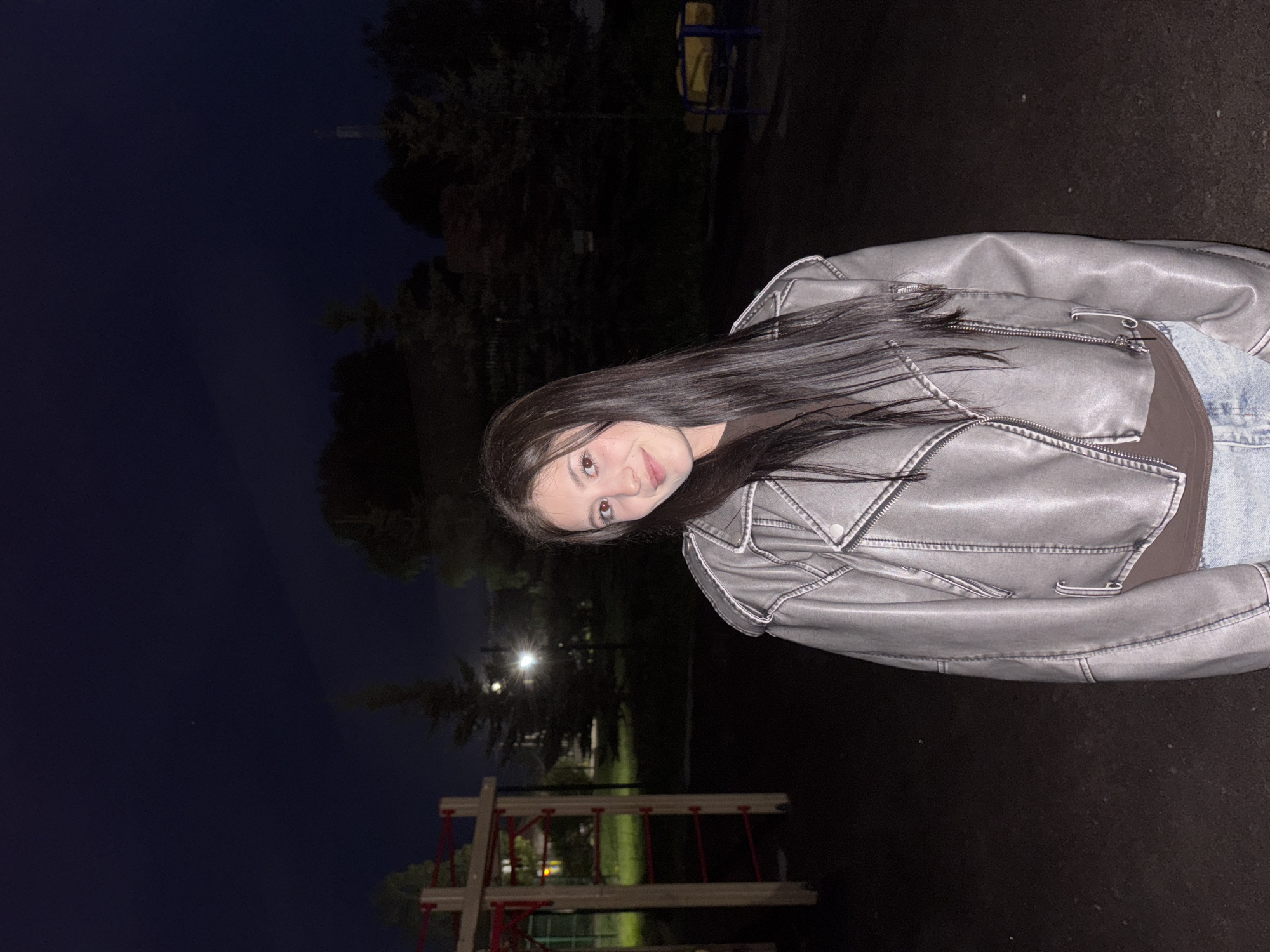 The image shows a young woman in a gray jacket, standing in a park in Фоки, Russia at night. The background features trees, playground equipment with red accents, and a dark sky. The woman is facing the camera. The scene appears to be illuminated by a streetlight, which is also visible in the background.