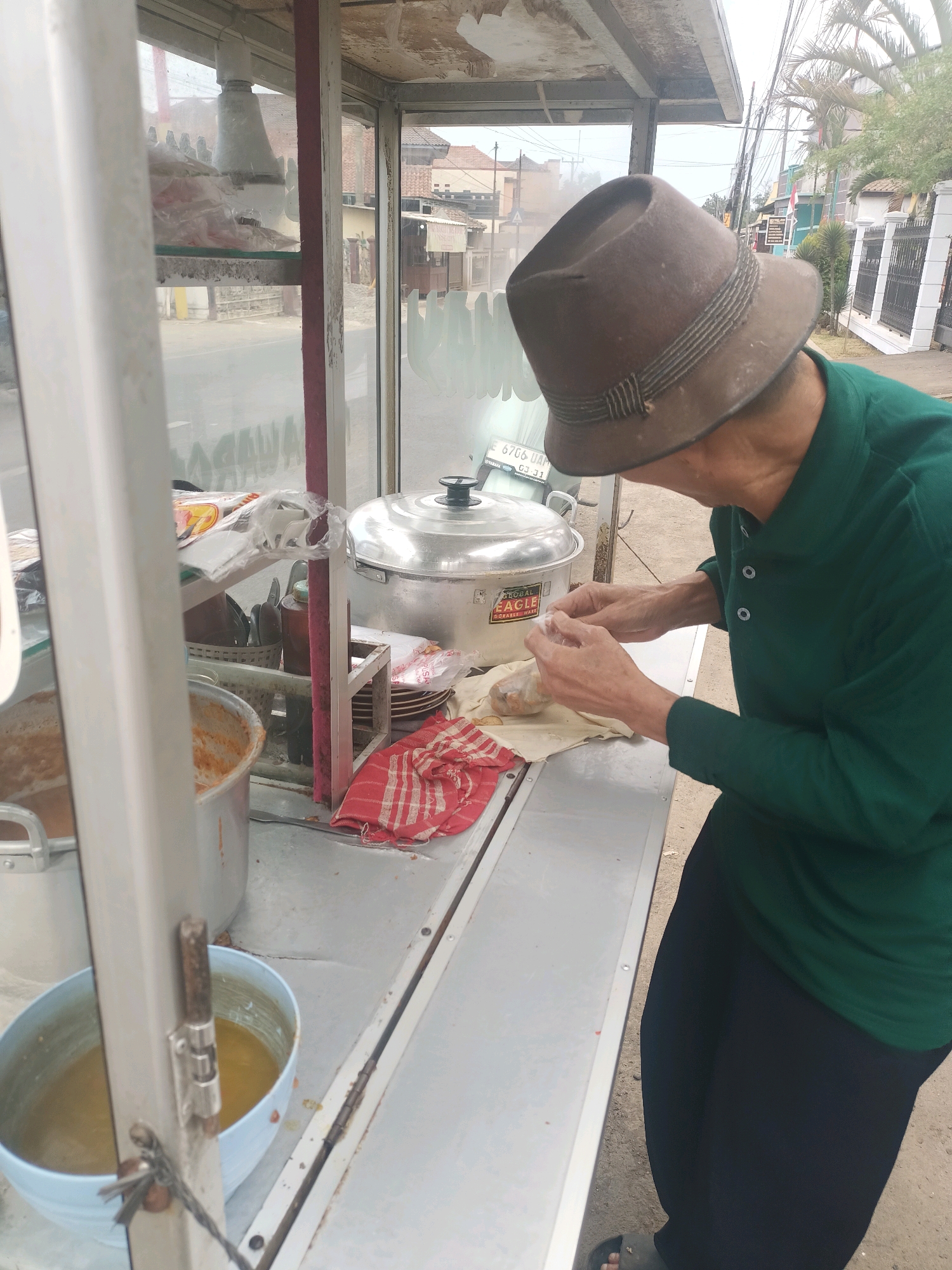 In Sukamukti, Indonesia, an individual wearing a brown fedora and a green long-sleeved shirt is preparing food at a street food stall. The stall features a silver countertop, a large aluminum pot with a black lid, and various containers of food. Behind the stall, a street with houses and a fence is visible. The person's hands are actively engaged in wrapping food, suggesting food preparation or packaging. The overall scene depicts a common urban street food vendor operation.