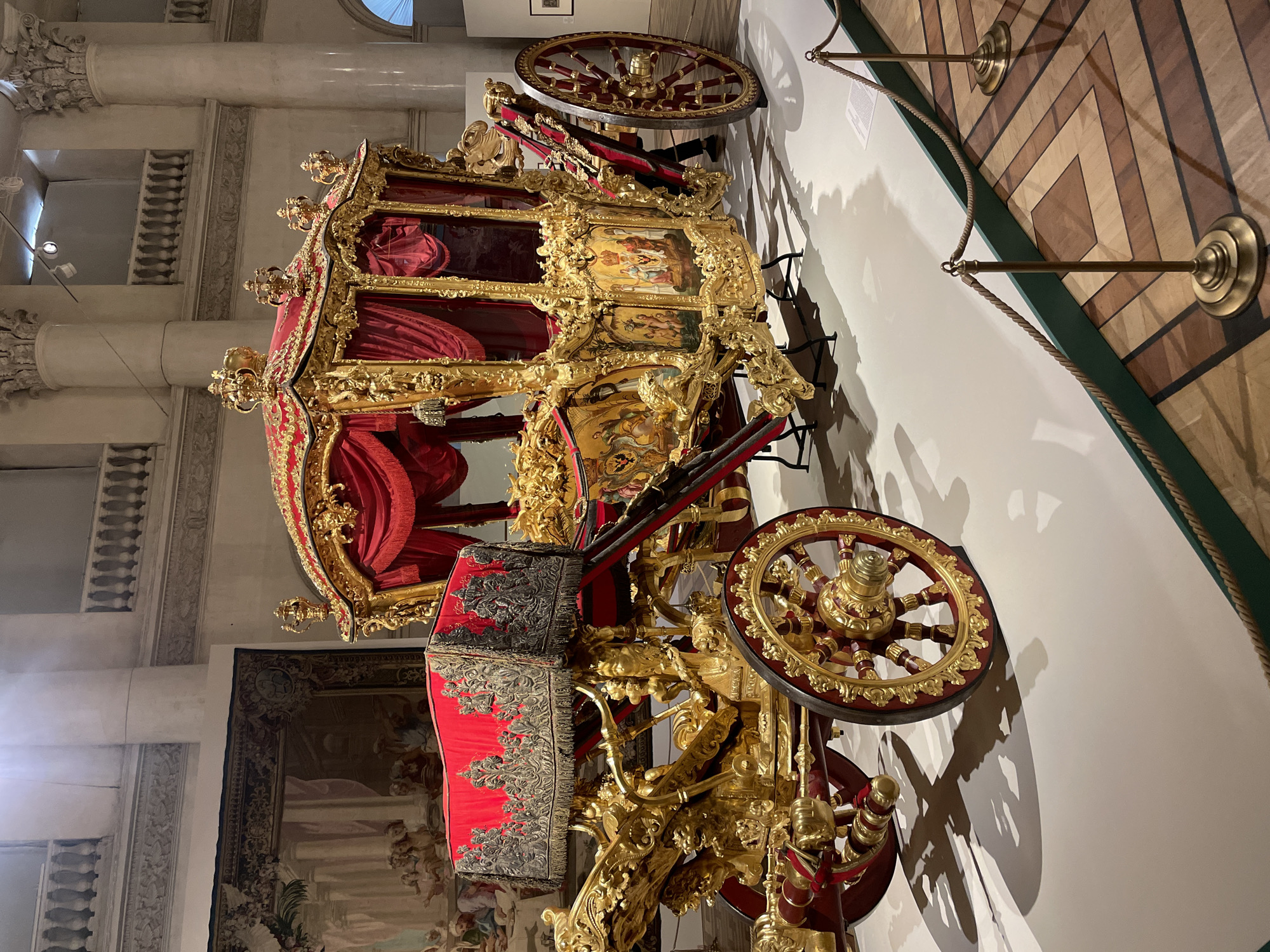 The image shows a large, ornate, golden carriage with red velvet interior and exterior trim. The carriage is on display indoors, likely in a museum or historical building. The location is identified as Saint Petersburg, Russia, based on the architectural features of the interior, including the columns and the tapestry on the wall. The carriage is surrounded by a rope barrier. The floor is a parquet pattern. The image appears to be taken at a low angle.