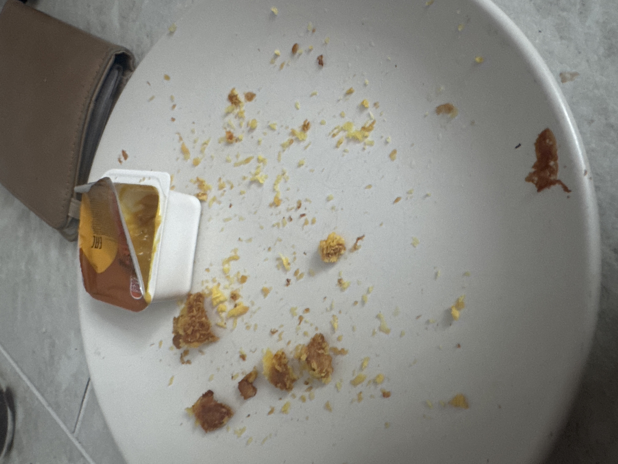 The image shows a white plate on a light gray tiled surface. On the plate, there are remnants of food, appearing to be breaded chicken pieces and crumbs, along with a small white container of dipping sauce, likely mustard or honey mustard, with the lid partially peeled back. The plate is not clean, indicating that a meal has been consumed. In the background, a tan leather wallet or clutch is visible, placed on the tiled surface. The lighting suggests an indoor environment, possibly a home or a casual dining establishment. There are no people visible in the image. The scene is static and depicts the aftermath of a meal. No specific landmarks or indications of Saratov, Russia, are present in the image.