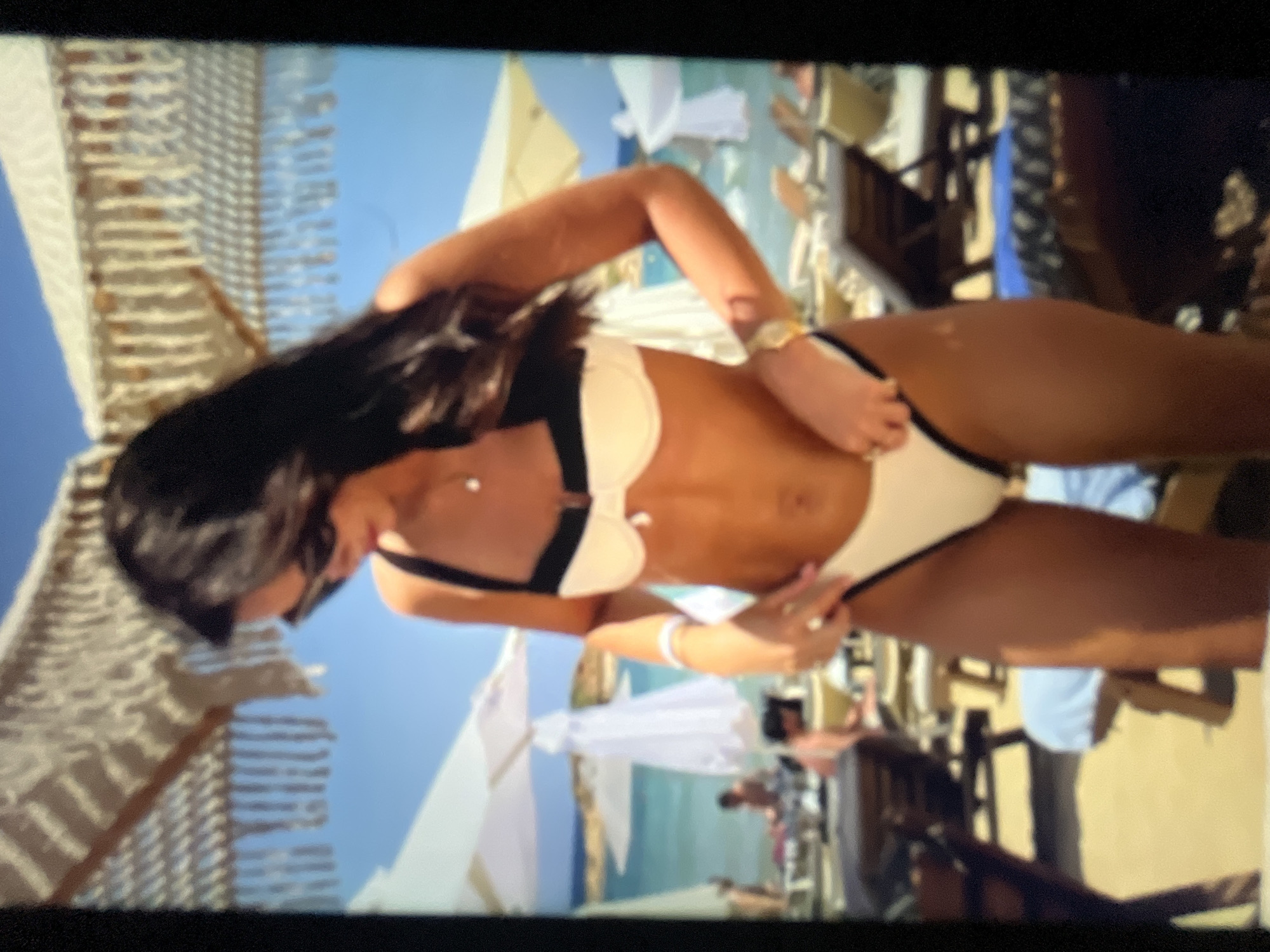 The image depicts a woman in a black and white bikini on a beach. She is wearing sunglasses and a necklace. Her hands are at her hips, with one hand appearing to adjust her bikini bottoms. In the background, there are beach umbrellas, lounge chairs, and the sea. There are also other people in the background, some of whom are seated or walking. The scene suggests a sunny day at a beach resort. The location of the image is not specified as Sofia, Bulgaria, and there are no elements in the image to suggest this location.