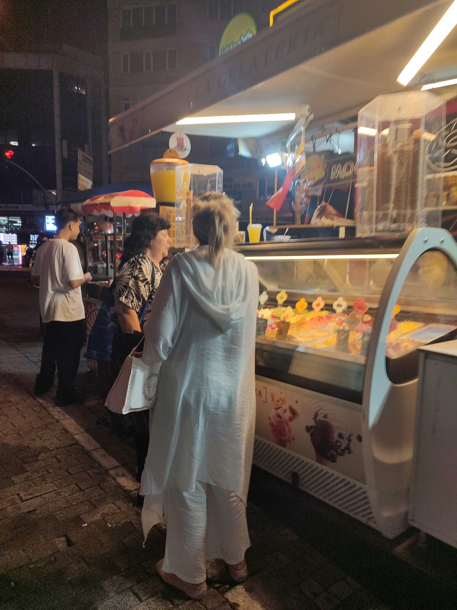 In Kadıköy, Turkey, at night, people are gathered around an outdoor gelato stand. A young man in a white t-shirt and black pants stands to the left, facing the gelato stand. To his right, a woman with dark hair, wearing a patterned shirt, is looking towards the gelato display. In the foreground, a woman in a white hooded outfit and white pants faces away from the camera, looking at the gelato. The gelato stand has a glass display case filled with various colorful gelato flavors. An umbrella with red and white stripes is visible behind the stand, and a large dispenser with orange liquid is positioned above it. The scene takes place on a cobblestone sidewalk in front of a building. The lighting is from the gelato stand's illuminated interior and overhead lights.