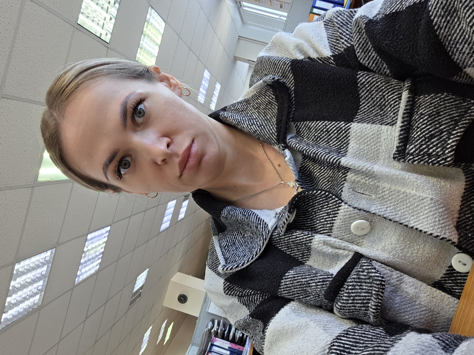 A woman with fair skin and light brown hair pulled back is looking into the camera. She is wearing a gray and black plaid flannel shirt and a thin silver necklace with a cross pendant. The environment appears to be an office or library space with white tiled walls and fluorescent lights. A cabinet with a clock and shelves with binders can be seen in the background. It is unknown if this image was taken in the city of Chaikovsky, Russia.