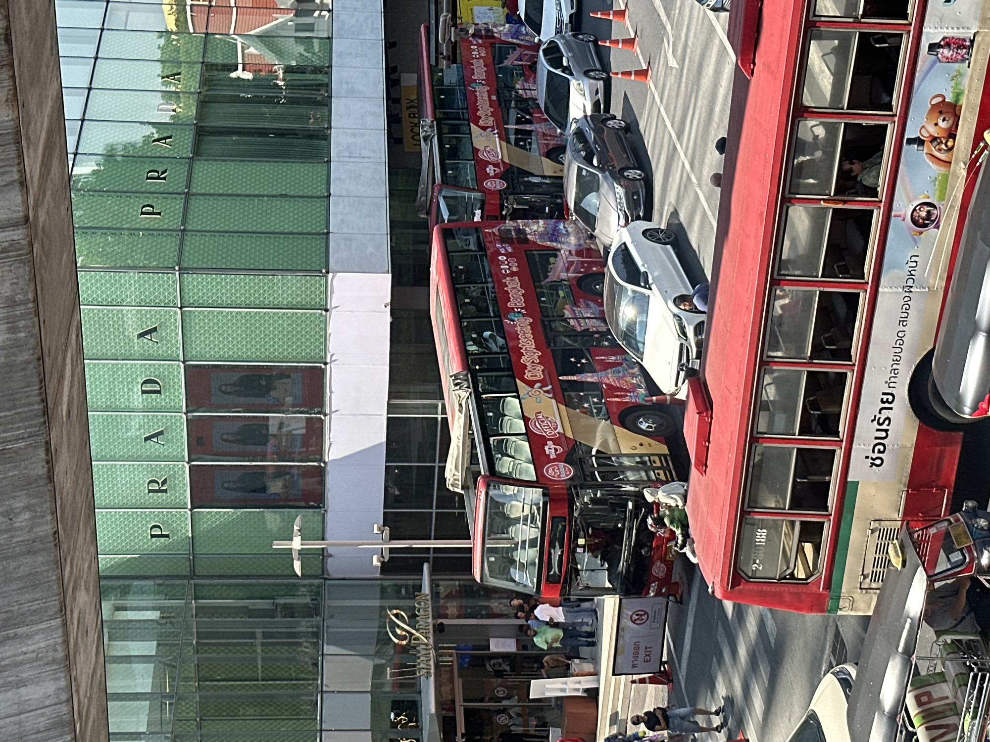 The image depicts a street scene in Bangkok, Thailand, near the Siam Paragon shopping mall. Prada signage is prominently displayed on the building's facade. Several red double-decker "City Sightseeing Bangkok" buses are visible, lined up on the street adjacent to the Prada store. People are seen in the vicinity of the buses and the mall entrance, including pedestrians walking and standing. Various cars and traffic cones are also present. A sign with the words "ทางออก EXIT" (Exit) is visible near the mall entrance.