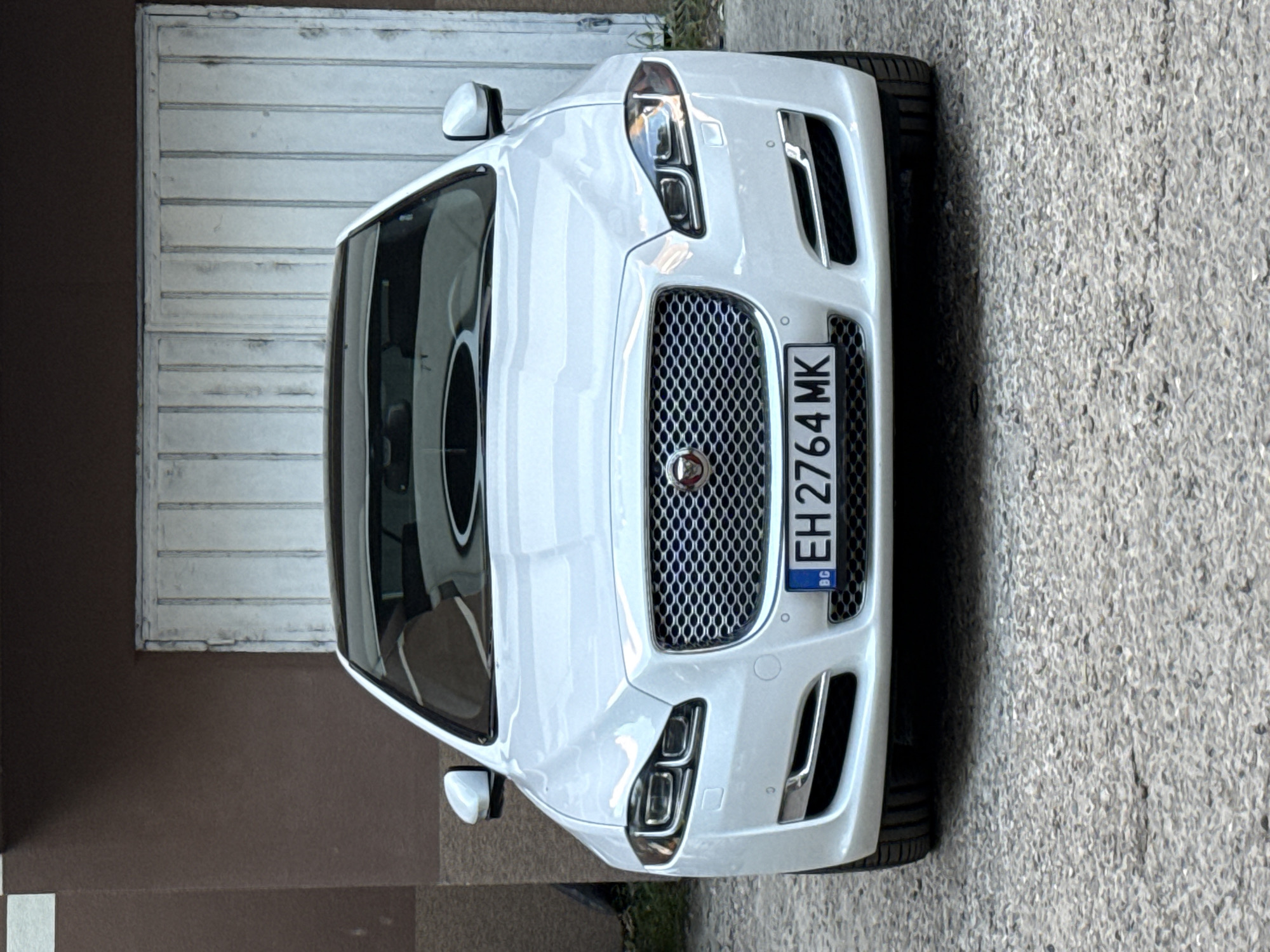 A white Jaguar car is parked on a concrete surface in front of a building. The car's license plate reads "EH 2764 MK." The building has a brown lower section and a white garage-style door with horizontal panels. The location is likely in Pleven, Bulgaria, based on the license plate which shows that the car is registered there.
