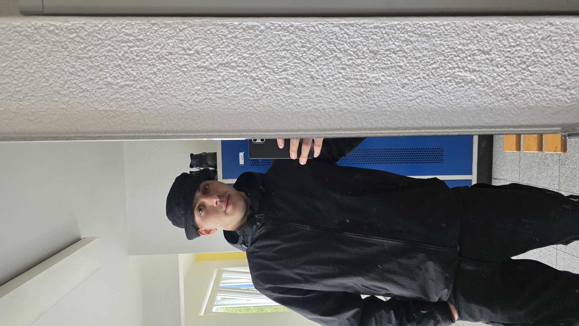 A man, wearing a black bucket hat, a black jacket, and black pants, stands in front of a mirror, taking a selfie with his phone. Behind him, a blue locker is visible, along with two pairs of boots on top of it. To the left of the locker, a window allows natural light into the room, revealing some greenery outside. To the right of the locker, a textured wall is prominent. The floor is tiled, and a wooden bench with three seats is visible in the background. The image appears to be taken indoors, possibly in a locker room or similar facility.