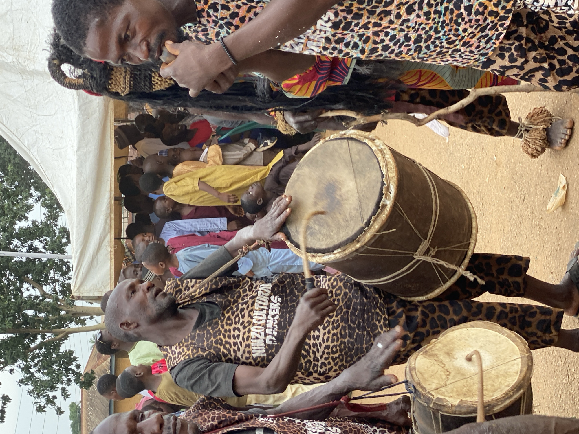 In Kafanchan, Nigeria, a gathering is observed, featuring a group of people with specific cultural characteristics. Two individuals in animal print attire are playing traditional drums. One person appears to be playing a flute or similar wind instrument. In the background, a crowd is visible under a white tent, with trees surrounding the scene. The foreground shows the dusty ground.