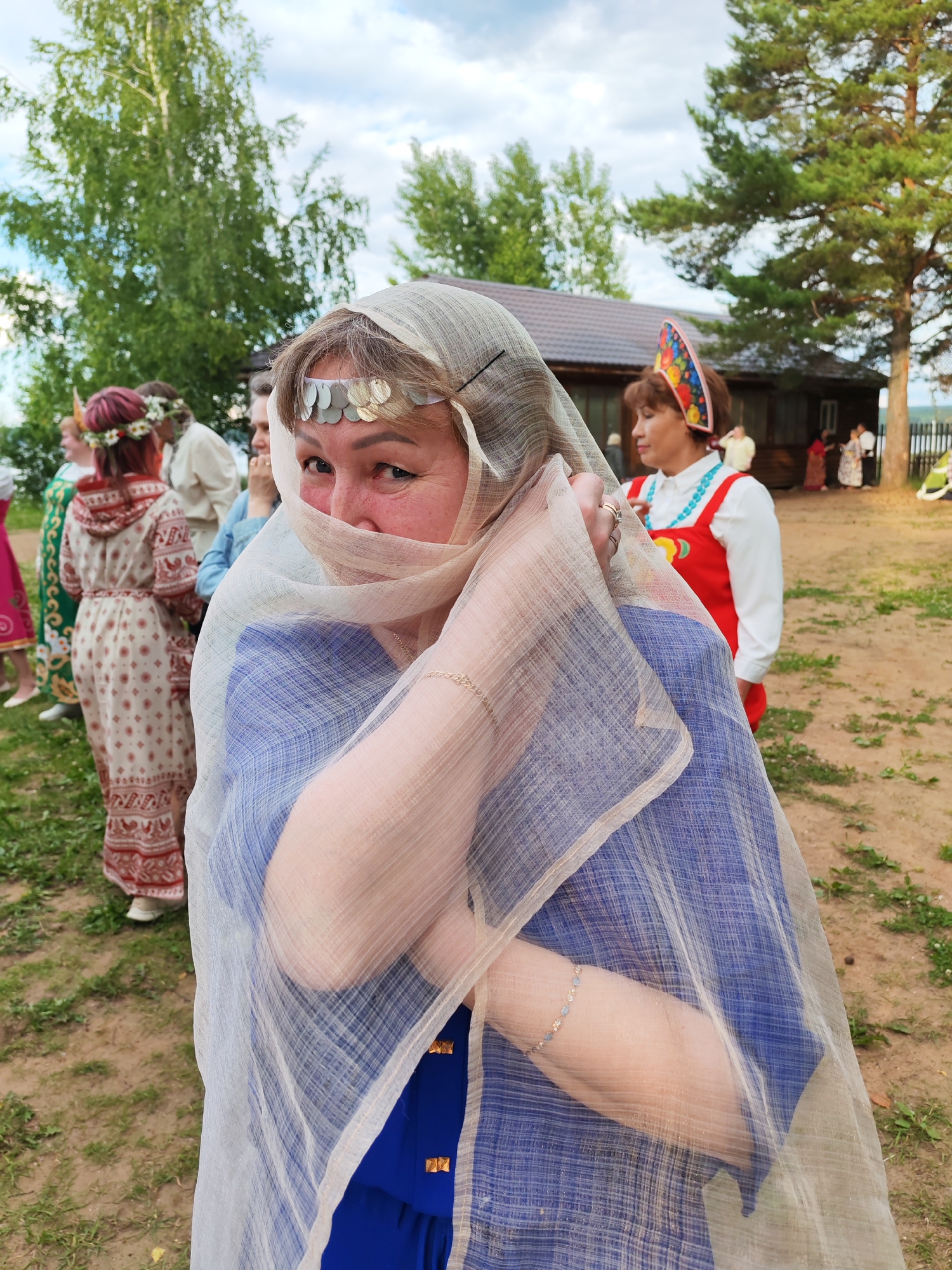 A woman in traditional dress, her face partially obscured by a sheer veil, looks directly at the camera. She wears a headpiece adorned with circular, metallic ornaments and a blue dress. The background shows other individuals in similarly styled attire, and a rustic wooden building, suggestive of a rural or historical setting. The scene appears to be outdoors with a cloudy sky and trees in the distance, possibly depicting a cultural festival or historical reenactment in or near Chaikovsky, Russia, a city known for its industrial and cultural heritage.