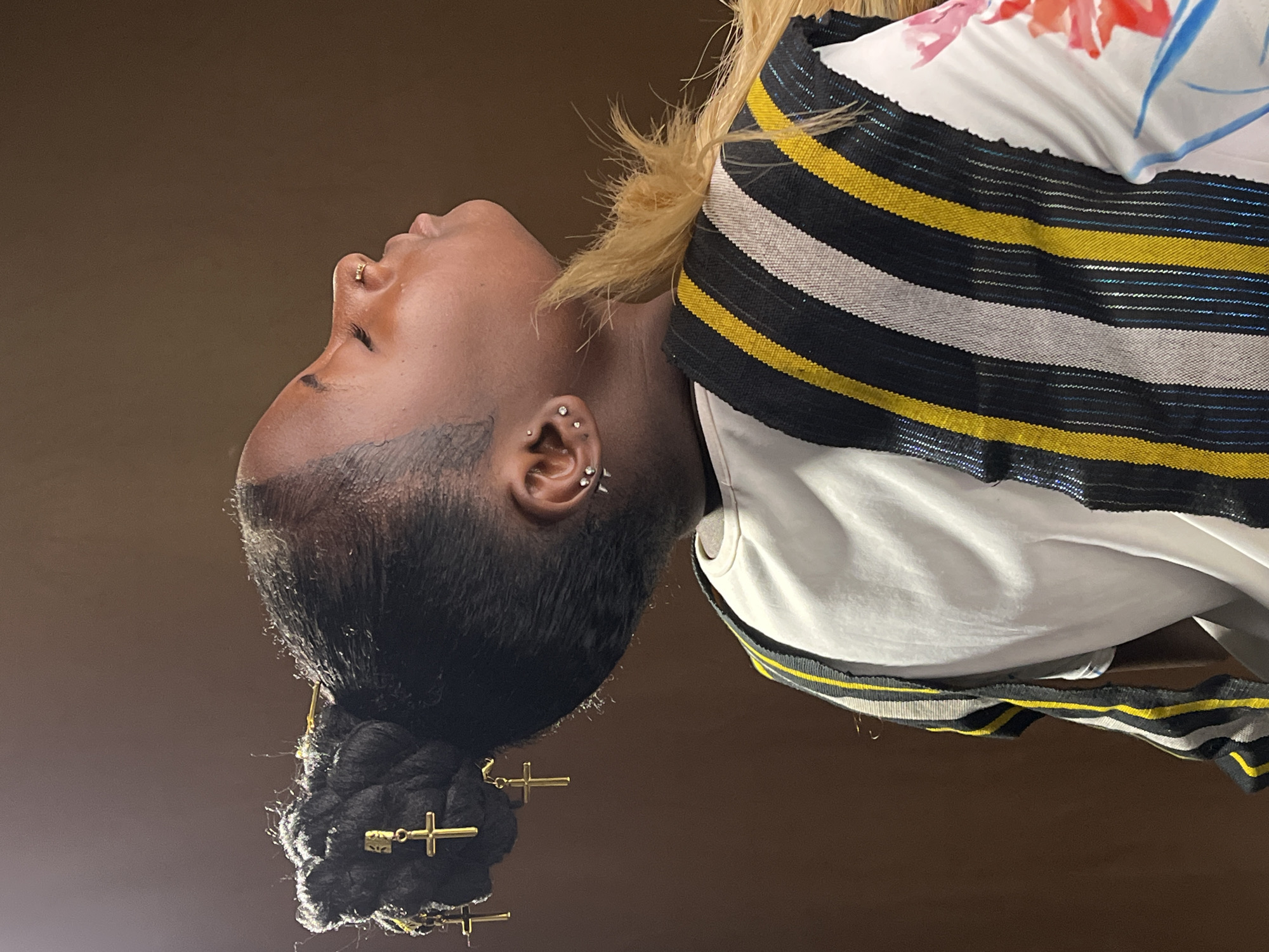 This close-up profile portrait, taken in Jos, Nigeria, features a person of dark complexion, positioned against a plain dark brown background. The subject's head is tilted upwards and slightly to the right, with eyes closed or looking downwards, conveying a tranquil or reflective mood.
The person's dark hair is meticulously styled into a bun or updo, embellished with several gold-colored hairpins designed as crosses and keys. A noticeable section of lighter, possibly blonde, hair is visible near the ear and neck, providing a stylistic contrast. Adornments include a small gold ring piercing on the nose and multiple ear piercings, comprising studs and small hoops.
The attire consists of a white garment, likely a top, with subtle hints of a colorful floral pattern in red and blue on the upper shoulder or back. Over this, a striking striped fabric is worn, featuring horizontal bands of black, yellow, and white, with some of the black stripes incorporating a blue, shimmery thread. The image focuses on the subject's personal style, intricate hair accessories, and jewelry. No text is visible in the media.
