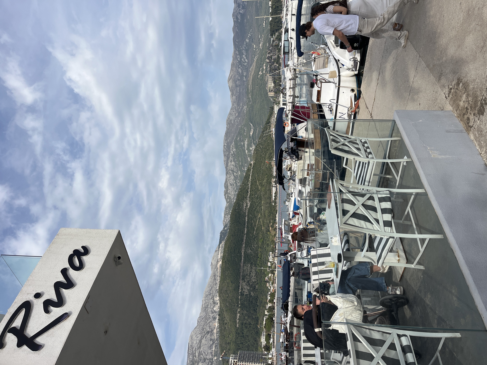 The scene shows an outdoor dining area with a view of a marina and mountains, likely located in Budva, Montenegro. A sign with the word "Riva" is prominently displayed on a building. Several people are present, including a person relaxing in a chair at a table, people walking along the dock. Various boats are docked in the marina. The sky is cloudy.