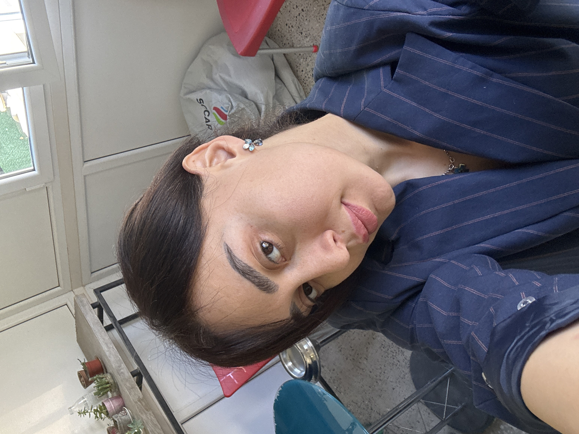 A woman is looking at the camera. The scene is indoors, possibly in an apartment in Tbilisi, Georgia. The woman has dark hair, brown eyes, and is wearing a navy blue pinstripe blazer with silver buttons. She has an earring in the shape of a flower and a delicate necklace. She is seated on a teal colored chair. In the background are various items, including a red stool, a white bag with the word "SCAR" visible, a window with a view to greenery, and a rack with small potted plants. The room has white walls. The angle of the photo suggests she might be holding the camera herself.