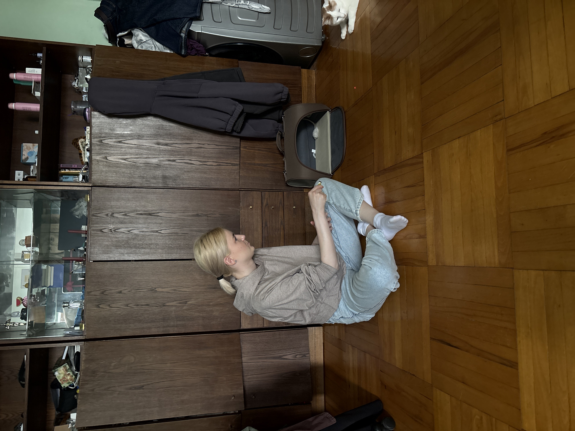 In Novoe Devyatkino, Russia, a young blonde woman wearing a light brown hoodie and light blue jeans sits on a wooden parquet floor. She is looking down at her hands, which are resting on her lap. To her right is a dark brown pet carrier with its door open. Further to the right, a white and brown cat is lying on the floor. Behind the woman and to her left is a large wooden cabinet with glass doors displaying various items. A dark gray hooded sweatshirt is hanging from the cabinet to the right of the carrier. A silver washing machine with clothes draped over it is visible to the far right of the image. The room appears to be a domestic setting.
