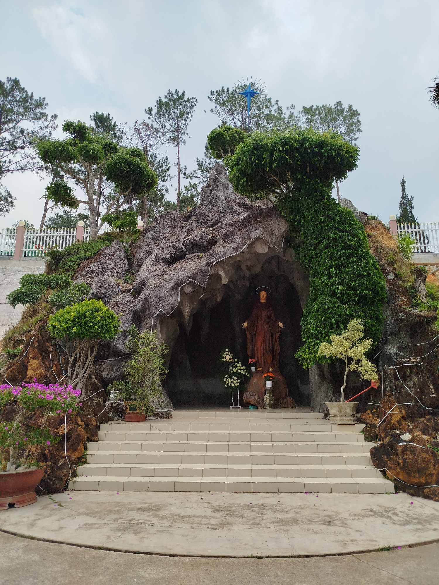 The image shows a Catholic grotto located in Lang Biang - Đà Lạt, Vietnam. The grotto is constructed from rock and is surrounded by trees and vegetation. A statue of a robed female figure with a halo is located inside the grotto, behind two vases of flowers. In front of the grotto are concrete steps leading up to a concrete platform. A white picket fence can be seen in the background. A blue star is positioned on top of the structure. Wiring is visible along the rock formations.