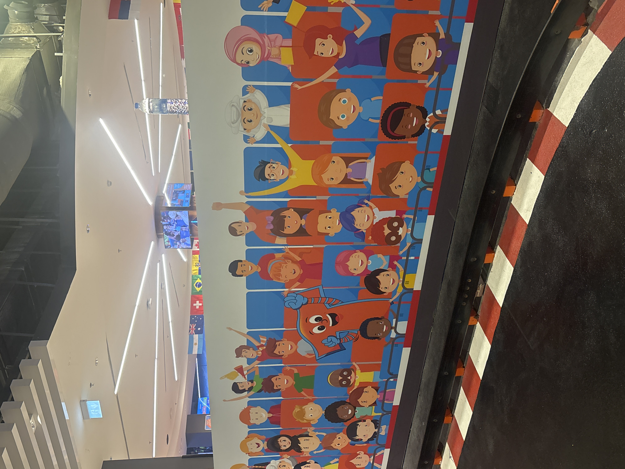 The image shows a colorful mural featuring cartoon children and adults in stadium seating, with a mascot character resembling the number seven in the center. The mural is positioned in front of a black barrier with red and white checkered markings, suggesting a race track or similar attraction. In the background, a ceiling with modern lighting and a few flags are visible, including the flags of Argentina, Australia, the United Kingdom, and Brazil. The overall scene suggests an indoor entertainment venue, potentially a theme park or a family-oriented attraction, and it is located in Dubai, United Arab Emirates.