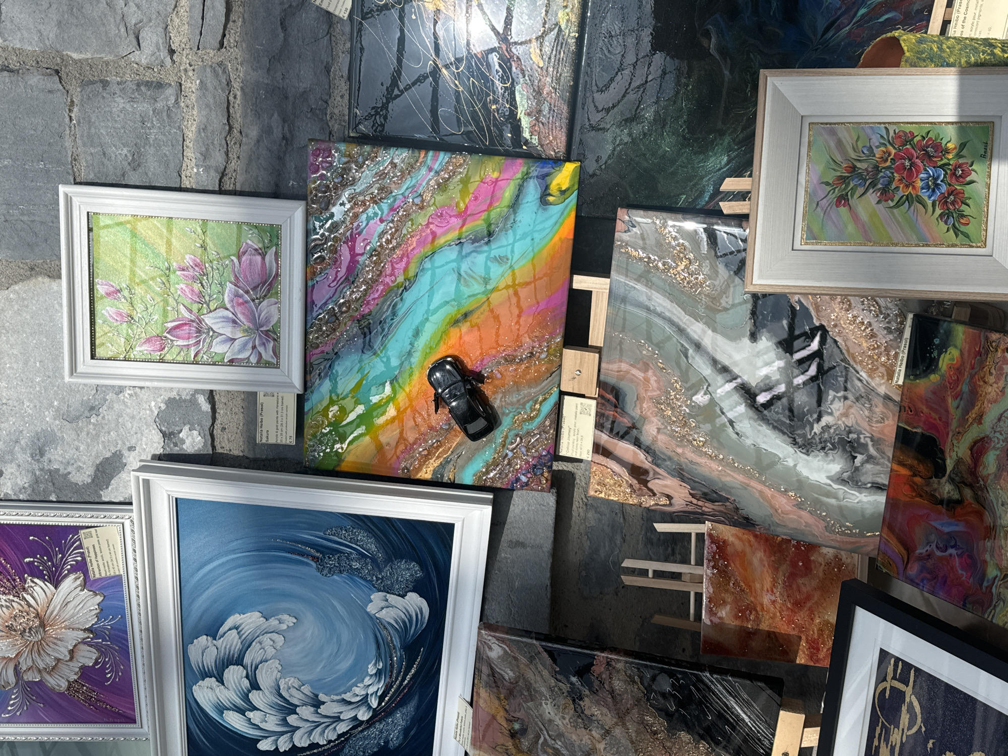 The image shows an outdoor art display against a stone wall in Kilkenny, Ireland. The display features several framed and unframed artworks, including paintings and resin art pieces, in various sizes and styles. Some of the art pieces are displayed on easels. The sun is casting shadows on the stone wall and the artworks, indicating it is daytime. There are no people visible in the image. The overall scene suggests a market or exhibition of local artists' work.