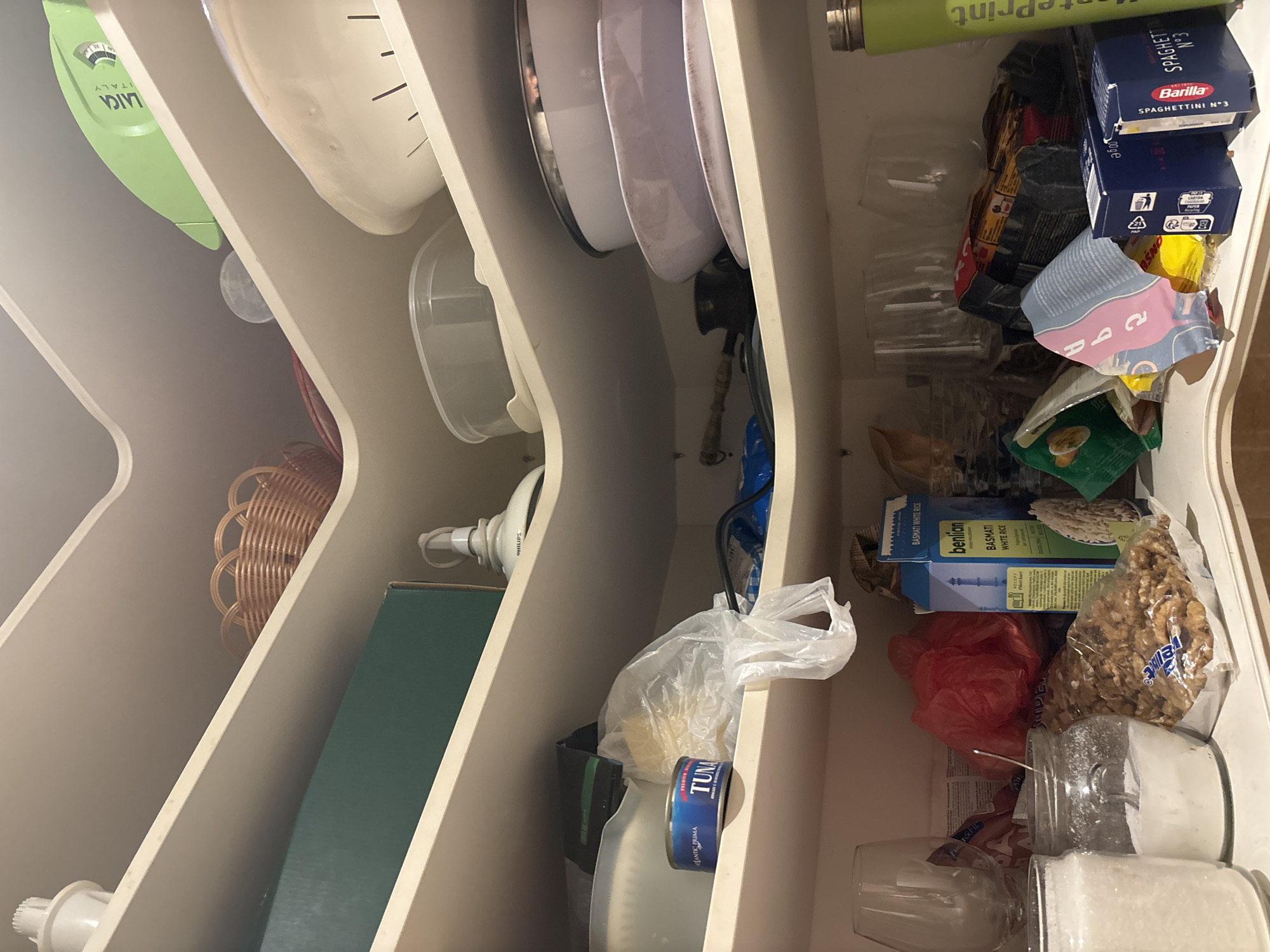 The image shows the inside of a pantry with multiple shelves filled with various food items and kitchenware. The pantry appears to be a built-in unit, possibly in a kitchen or dining area.
On the top shelf, a woven basket is visible, along with a green appliance and a large white bowl or colander. The middle shelf contains a green rectangular box, a white appliance with a cord, and a stack of plates and bowls. To the left of the plates, a blue can labeled "TUNA" is present, along with a plastic bag.
The bottom shelf is more cluttered and features several glasses, a jar filled with a white granular substance (likely sugar or salt), a bag of nuts, a red plastic bag, and several boxes of food items, including "BENILAN BASMATI WHITE RICE" and "Barilla SPAGHETTINI N°3". A green bottle with the label "MontePrint" is also visible on this shelf.
The overall impression is one of a well-stocked but somewhat disorganized pantry. The lighting suggests an indoor environment, possibly with artificial light. There are no people or animals present in the image. The context of Budva, Montenegro, is not directly evident from the visual content of the image itself.