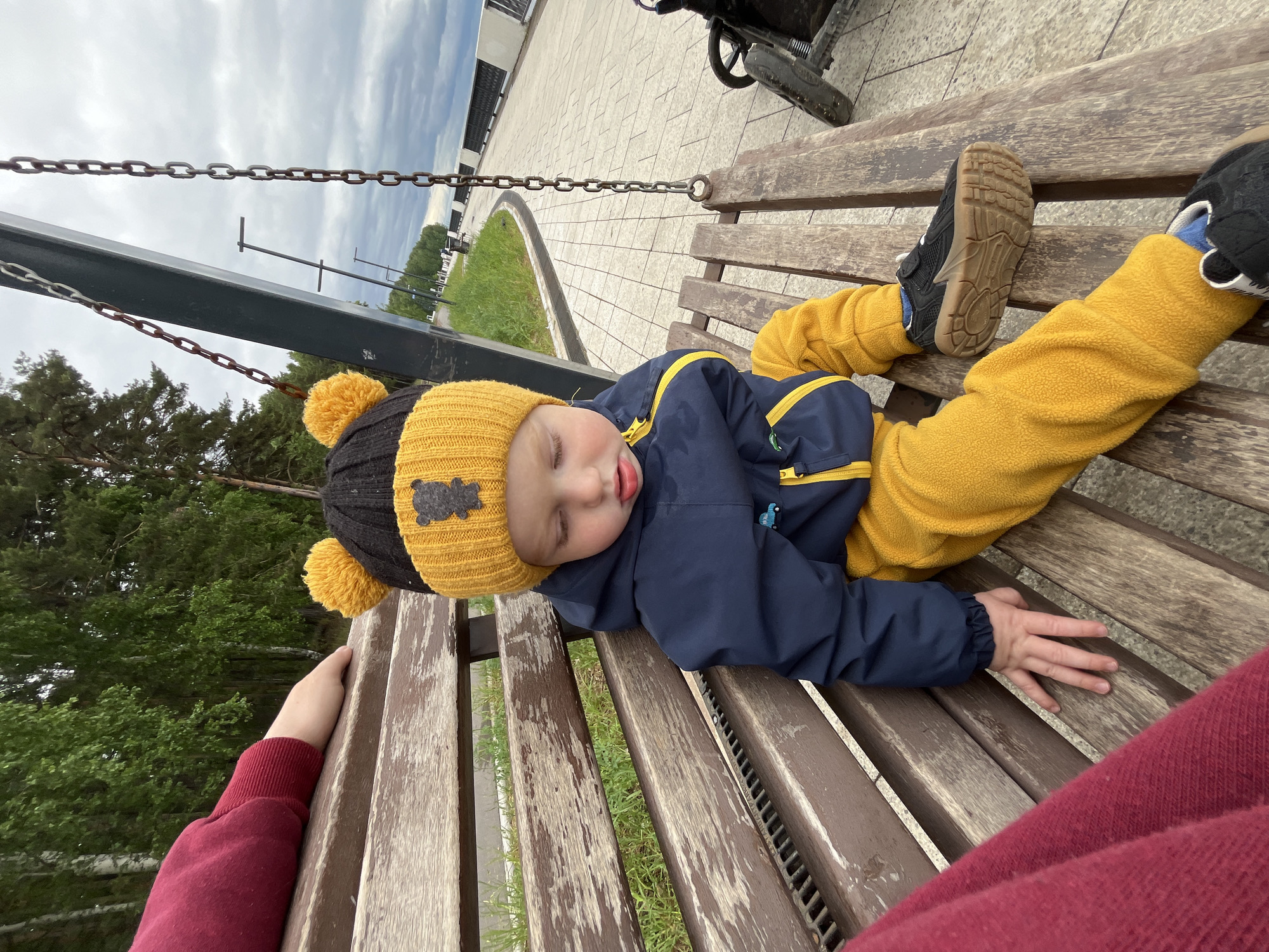 A young child wearing a black and yellow knit hat, a blue jacket with yellow trim, and yellow pants, lies asleep on a wooden park bench in Chaikovsky, Russia. The bench is suspended by metal chains. The child is wearing dark-colored shoes with light-colored soles. A person wearing a red sweater sits next to the child. Pavement is visible behind the bench, with grass and trees in the distance. The sky is overcast.