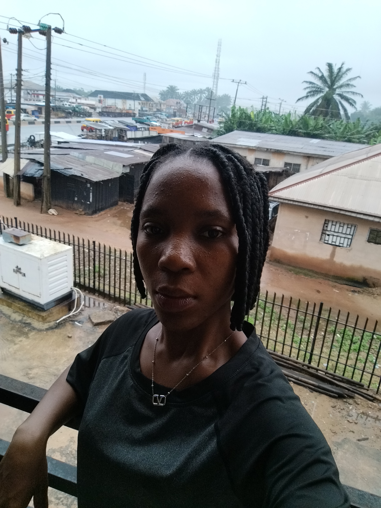 A woman with braided hair and a black shirt is shown from the chest up, looking directly at the camera. She is wearing a silver necklace. In the background, there are buildings, a generator, a fence, and a street with cars and trucks. The sky is overcast. This image was taken in Uselu, Nigeria.