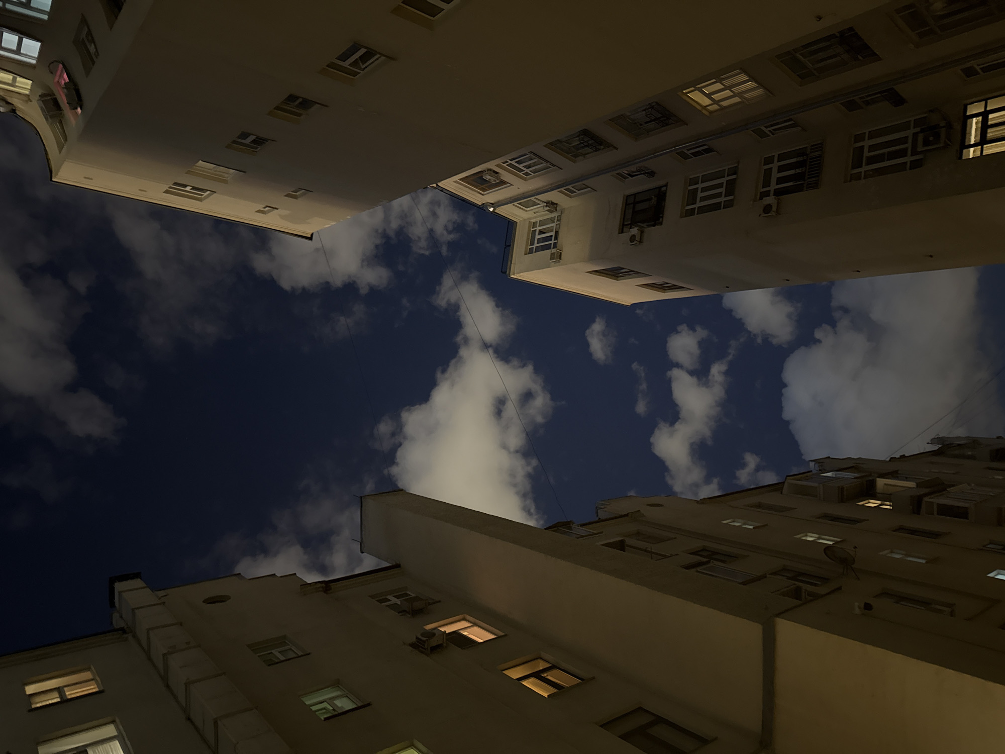The image presents a striking low-angle view looking upward from within a narrow urban courtyard, framed by tall, multi-story buildings. Three main building facades dominate the composition, appearing to converge towards the top of the frame, creating a sense of enclosure and scale.
The buildings themselves are constructed from a light-colored material, likely stucco or stone, and feature numerous rectangular windows. Many of these windows are illuminated from within, casting warm, yellowish light that contrasts with the dark exterior of the structures and the night sky. Details visible on the building exteriors include multiple window-mounted air conditioning units, particularly on the right and bottom buildings, and a small satellite dish affixed to a wall on the bottom-right building. One building on the left exhibits a large, vertical, rectangular duct or chimney-like structure running up its facade.
Above the buildings, the sky is a deep, dark blue, indicating nighttime. Scattered, irregularly shaped white clouds are visible against this dark backdrop, some appearing softly lit, likely reflecting ambient city light from below.
No people or discernible activity are present, though the lit windows imply habitation or use within the buildings. The image conveys a mood of quiet urban grandeur and observation from a constrained perspective. The time of day is clearly night, and the weather features scattered clouds. The scene is located in Moscow, Russia. No visible text appears in the image.