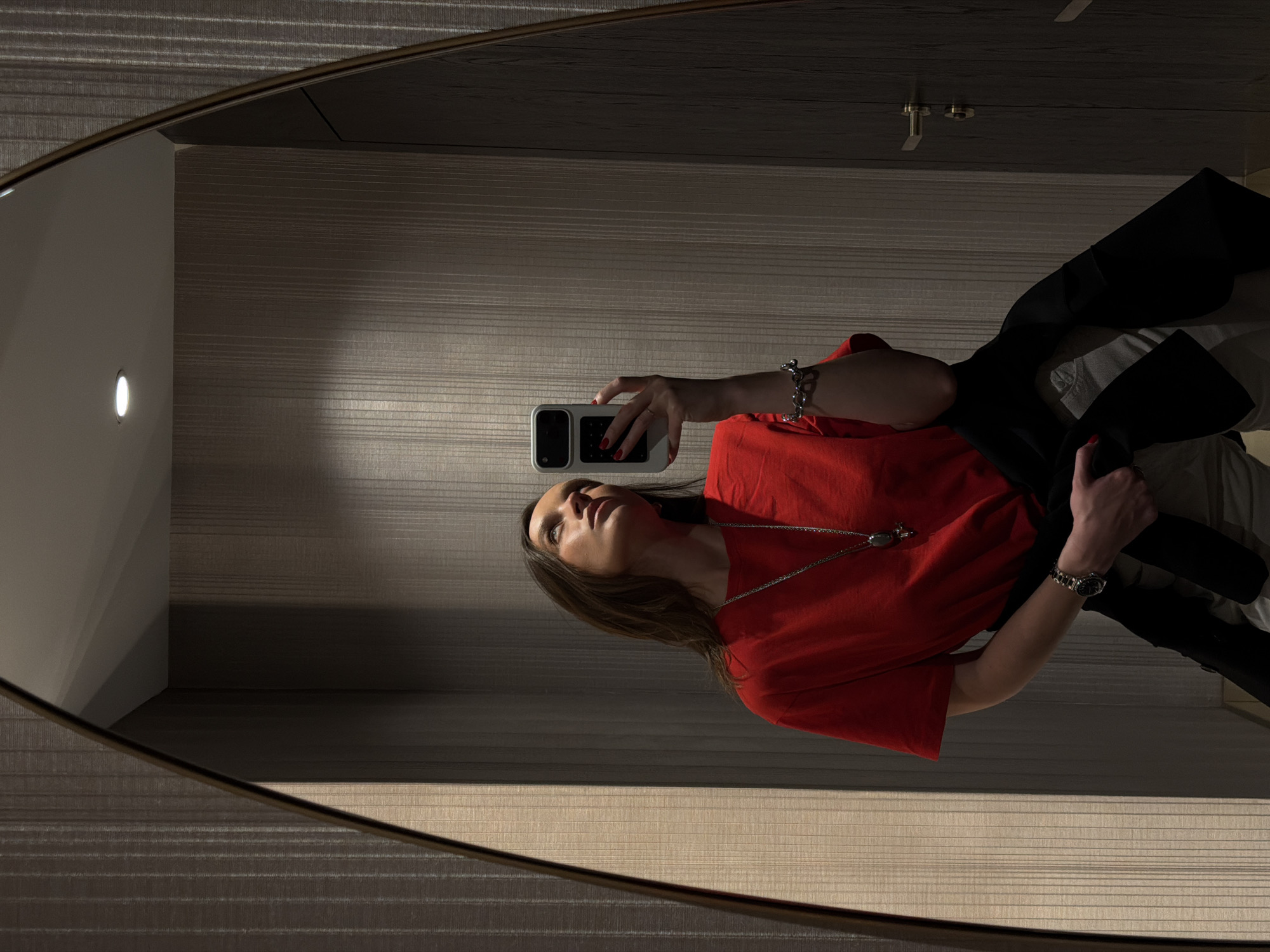 A female individual is depicted taking a mirror selfie in an indoor setting within the city of Budva, Montenegro. The subject has long, dark hair and is wearing a red short-sleeved top, a long silver chain necklace with a large pendant, a silver link bracelet on her left wrist, and a silver-toned wristwatch on her right wrist. Her fingernails are painted red. She holds a smartphone encased in white in her right hand, directing it towards her reflection. Her left hand holds a dark garment, possibly a jacket, which partially obscures light-colored trousers. Her gaze is directed upwards. The background, as observed in the oval-shaped mirror with a dark frame, consists of walls covered in vertically striped, light brown or grey wallpaper. A white ceiling with a single recessed downlight is visible. To the right, a dark wooden door with brass or gold-colored handle and lock hardware is present.