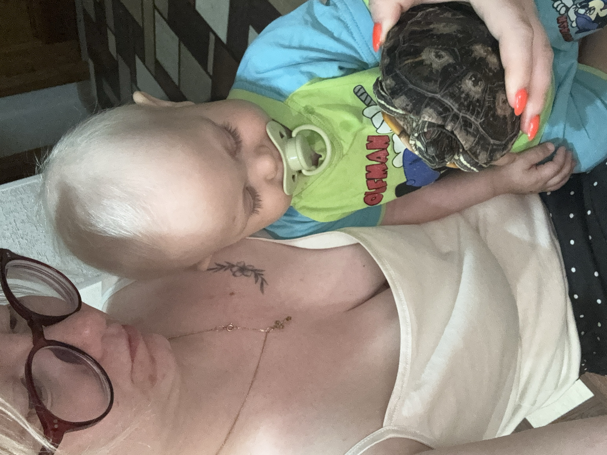 A close-up, vertical, waist-up shot shows a woman holding a baby. The woman, on the left, is wearing glasses and a white tank top. Her hair is blonde and pulled back. The baby, on the right, is looking down at a turtle in its hands and has a pacifier in its mouth. The baby is wearing a blue and green shirt. The turtle is dark brown with intricate patterns on its shell. The background is blurred but appears to be an indoor setting with a patterned floor. No specific location in Chaikovsky, Russia is identifiable.