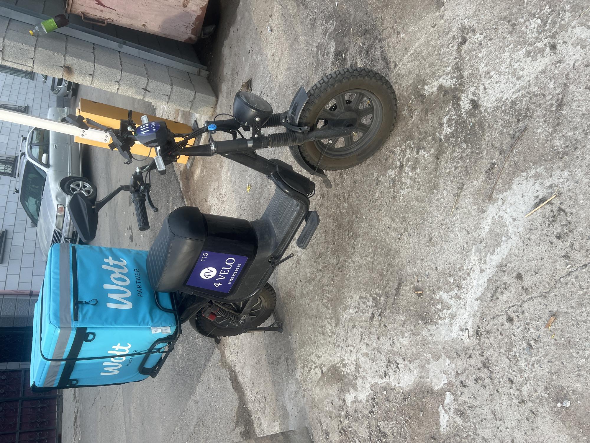 An electric scooter with a blue "Wolt" delivery bag is parked on a cracked concrete surface in an urban environment. The scooter is black and has a license plate with "4 VELO" and a phone number. A yellow traffic barrier stands behind the scooter. In the background, a silver car and a building are visible. The scene appears to be a street or alleyway in the city of Almaty, Kazakhstan.