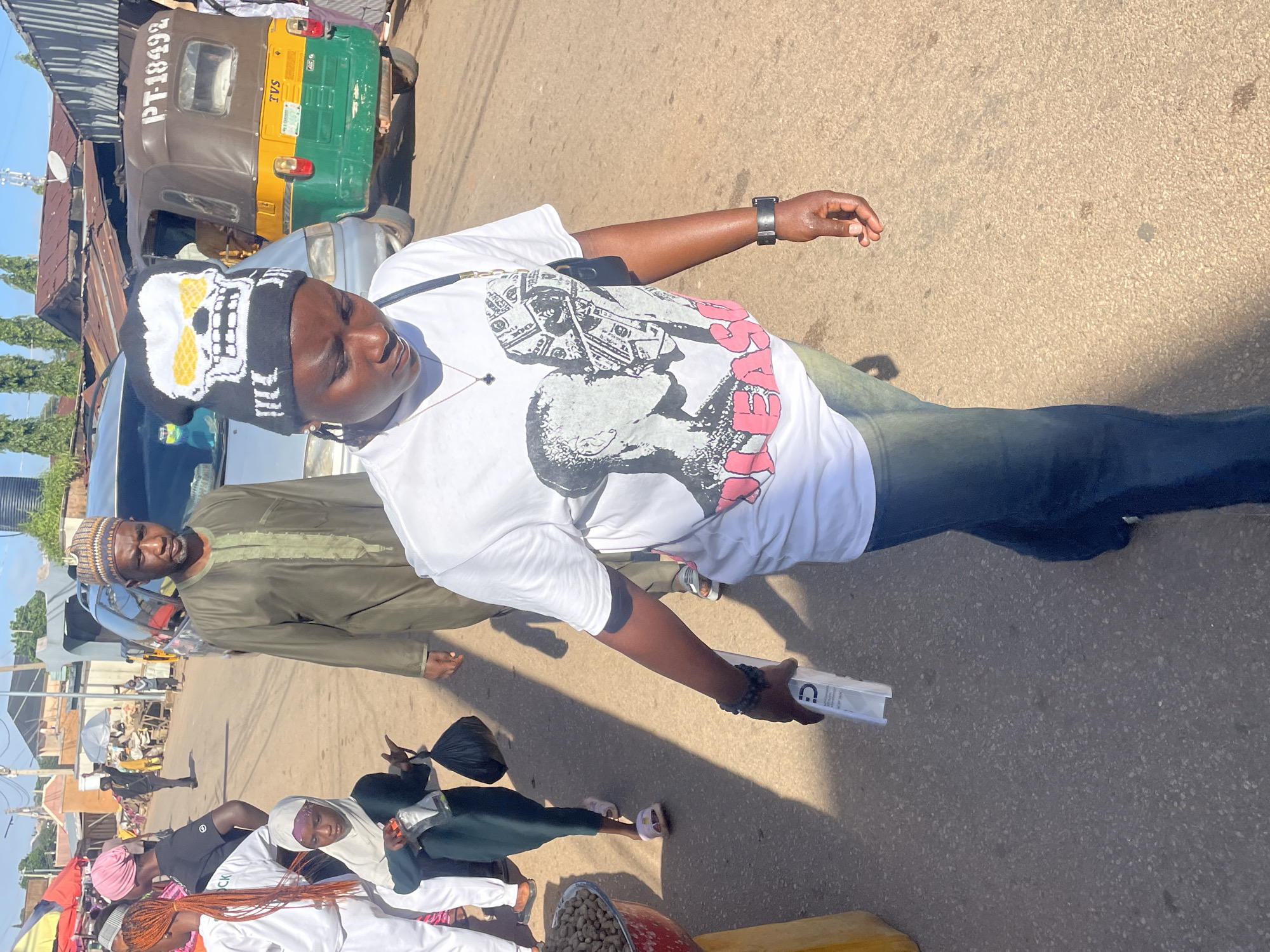 The image depicts a street scene in Jos, Nigeria, likely a market area due to the presence of vendors and various vehicles. In the foreground, a woman wearing a white t-shirt with a graphic print and blue jeans walks towards the viewer, holding a white rectangular object and wearing a skull-patterned beanie. To her left, a young girl in a green dress and a white hijab walks away from the viewer, carrying a black plastic bag and a transparent bottle. Behind them, an older man in a traditional olive green robe and a patterned cap walks away from the viewer. In the background, a green and yellow auto-rickshaw, identified by the license plate "PT-18492", is parked. Other people, market stalls with umbrellas, and buildings are visible further down the street. The ground is paved and appears somewhat uneven. The overall atmosphere suggests a busy, outdoor marketplace.